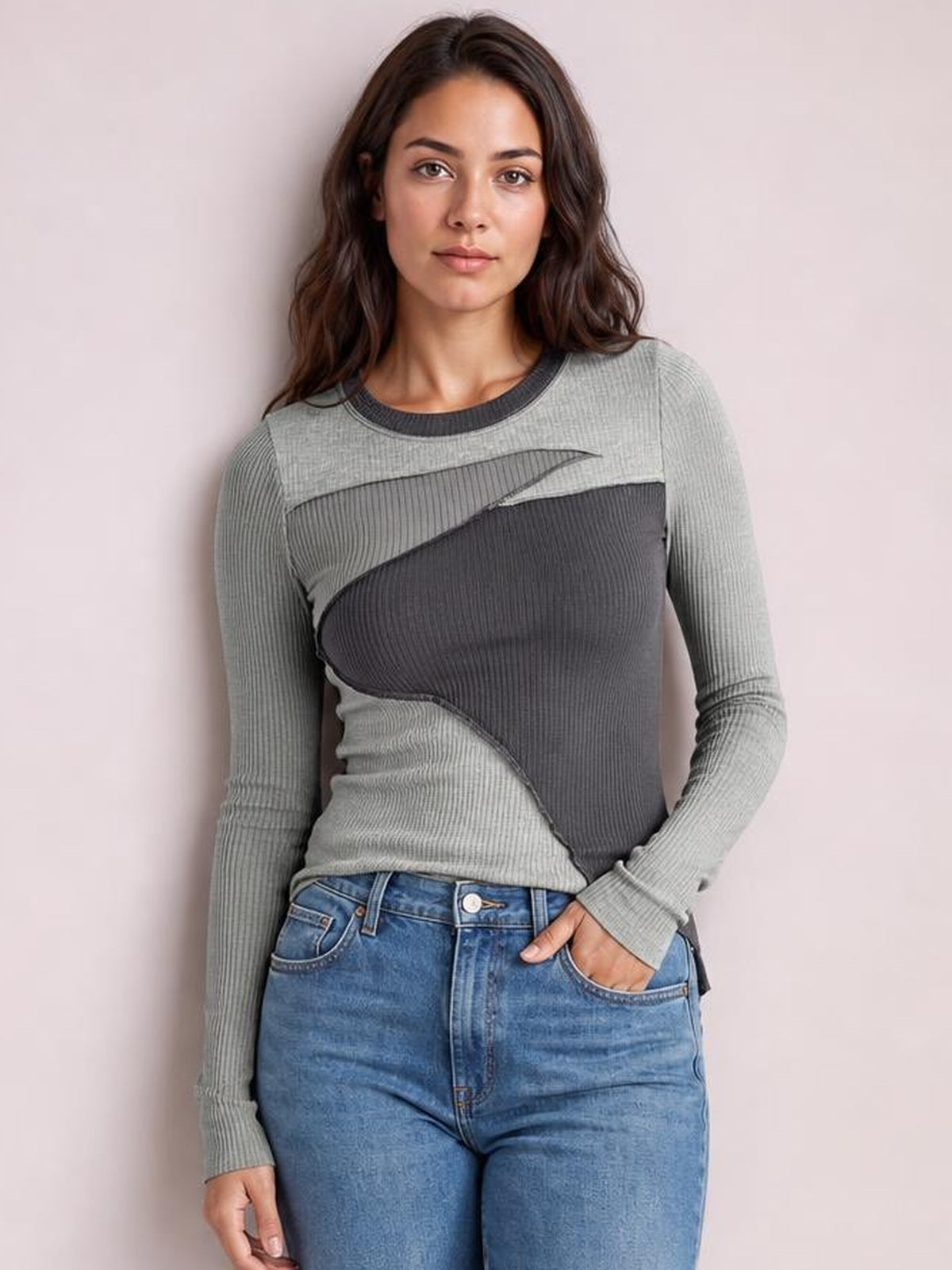 Woman wearing a gray contrast round neck long sleeve T-shirt with exposed diagonal seams and a fitted ribbed silhouette, posing with one hand in her jeans pocket.