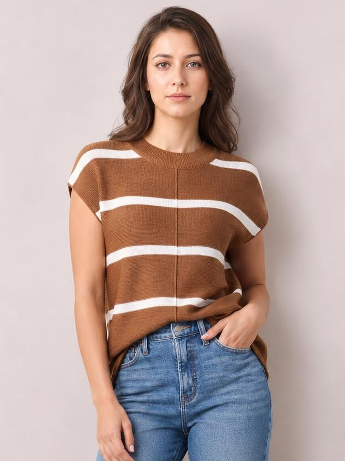 A model wears a brown knit top with contrasting white horizontal stripes, round neck, exposed center seam, and a relaxed short-sleeve silhouette while posing with one hand in her jeans pocket.