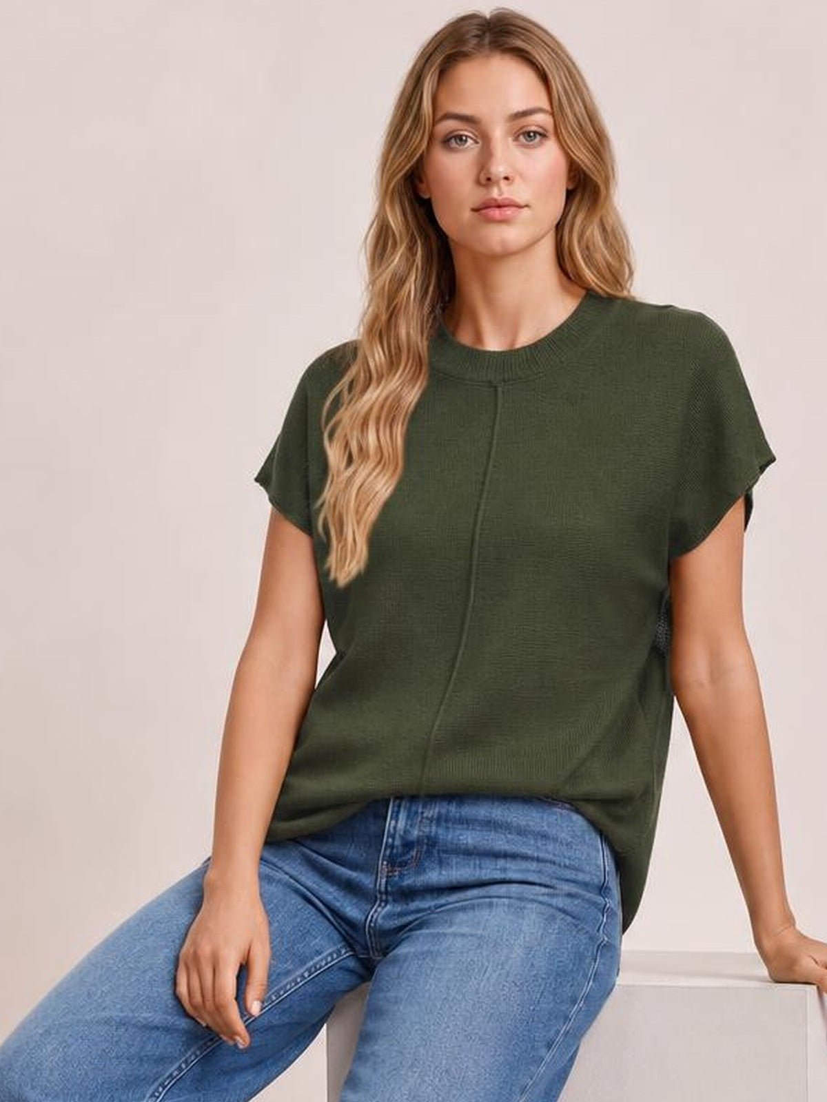Model seated in a dark olive green short-sleeve sweater with a round neck, exposed center seam, relaxed silhouette, and slightly stretchy knit construction.