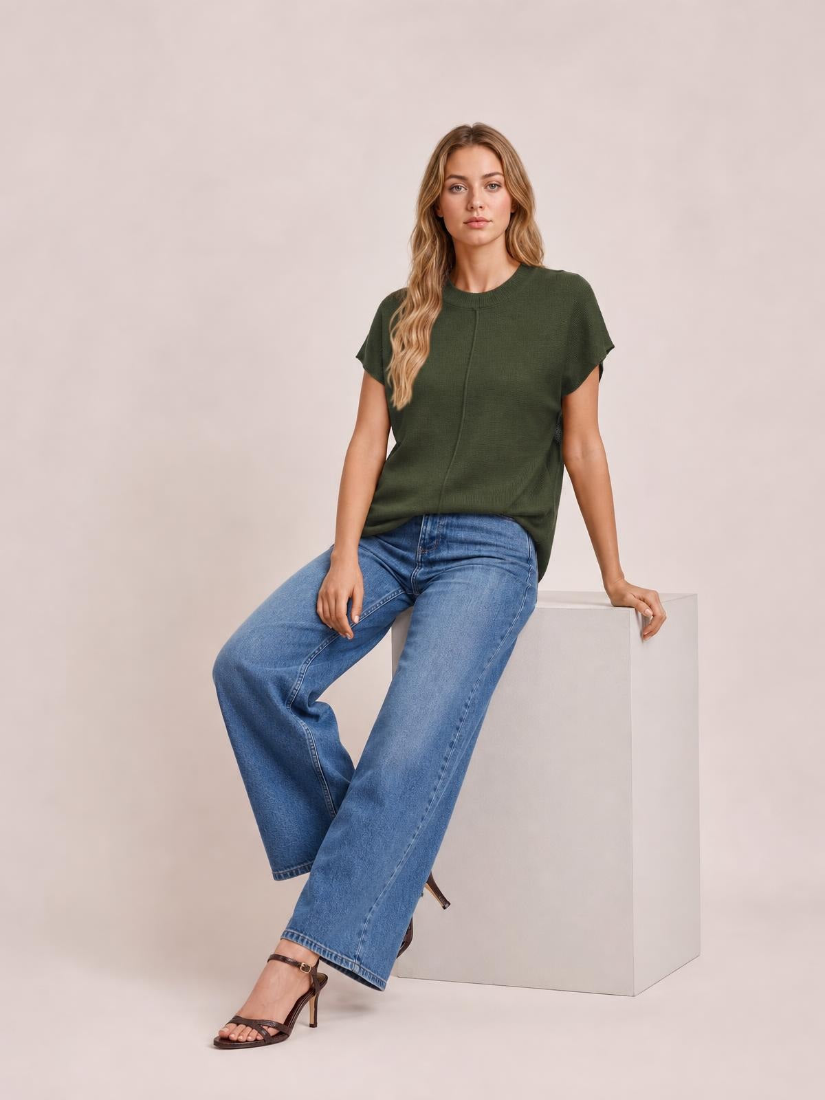 A model sits wearing an olive green round-neck short-sleeve sweater with an exposed center seam and relaxed silhouette.