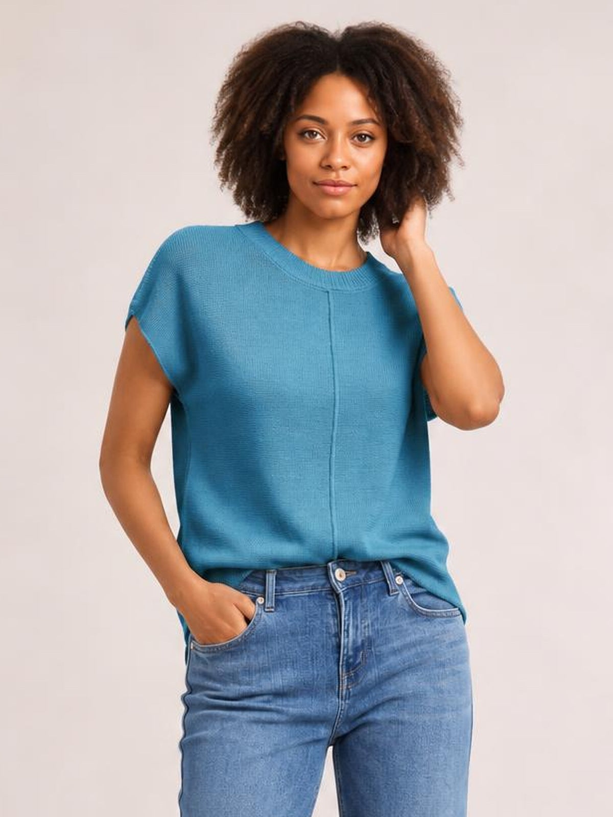 Model poses in a blue exposed-seam round-neck short-sleeve sweater with a relaxed straight silhouette, one hand in her jeans pocket and the other touching her hair.