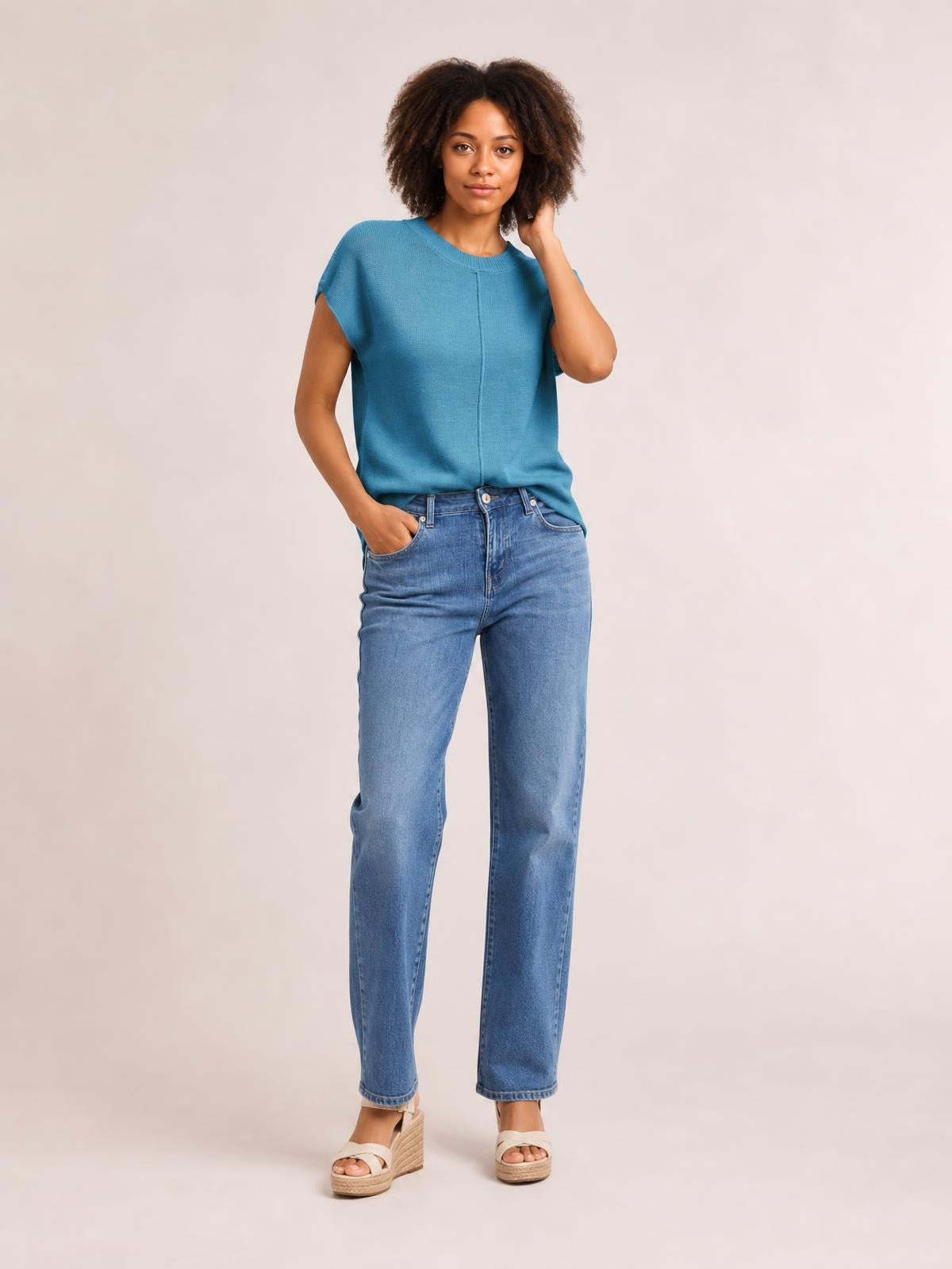 Model wearing a blue round-neck short-sleeve sweater with exposed center seam detailing and a relaxed straight silhouette, posing with one hand in her jeans pocket.