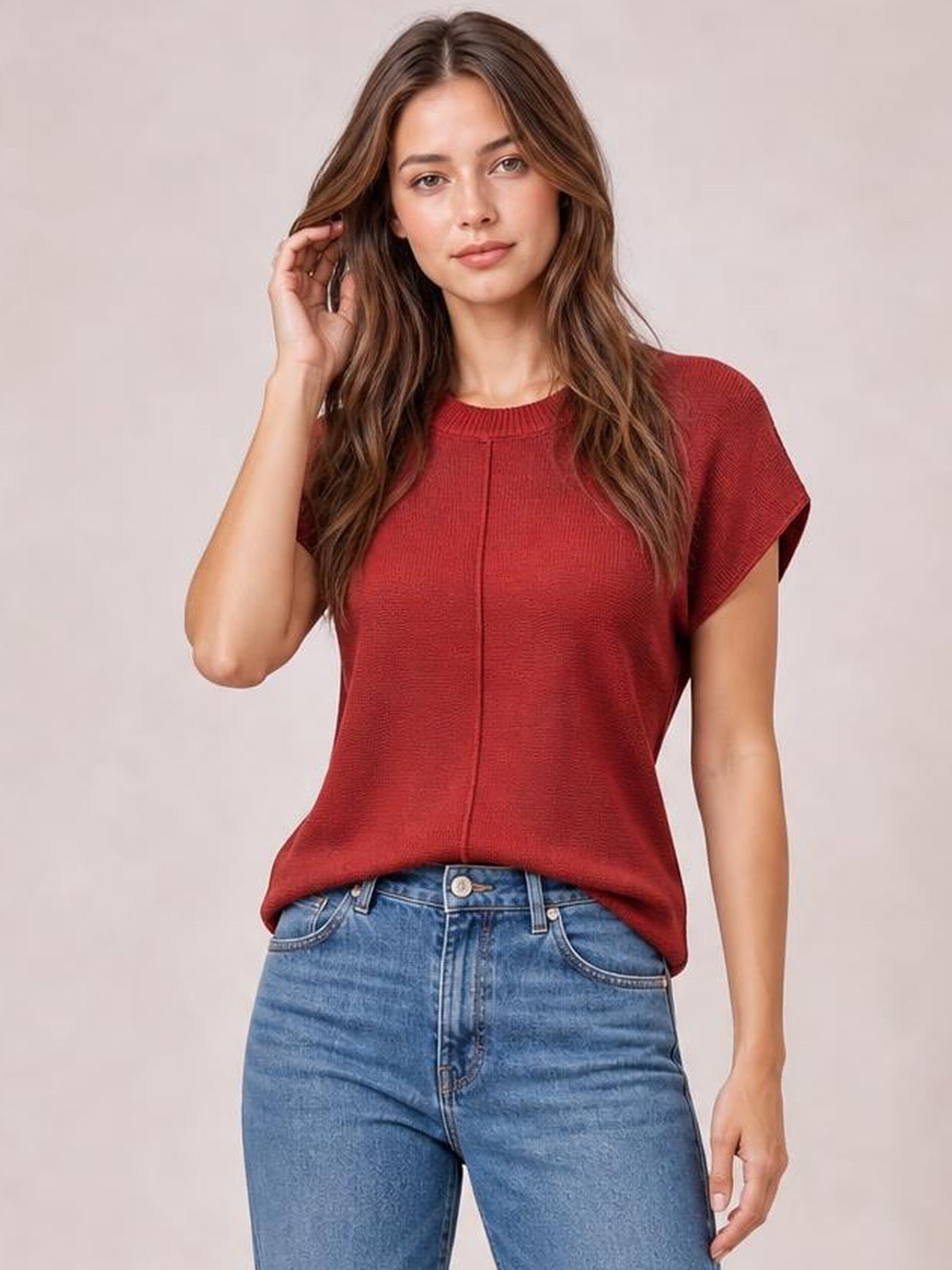 A model poses in a rust-red round neck short sleeve sweater with exposed center seams and a relaxed silhouette, touching her hair.