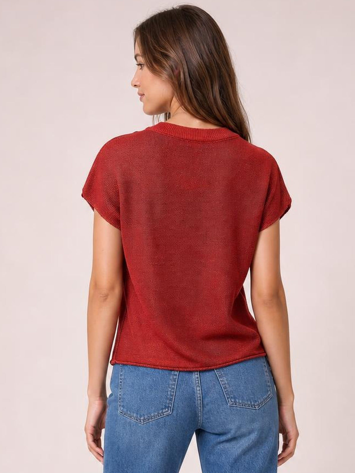 A model poses from the back in a red exposed-seam sweater with a round neck, short sleeves, and a relaxed straight silhouette.