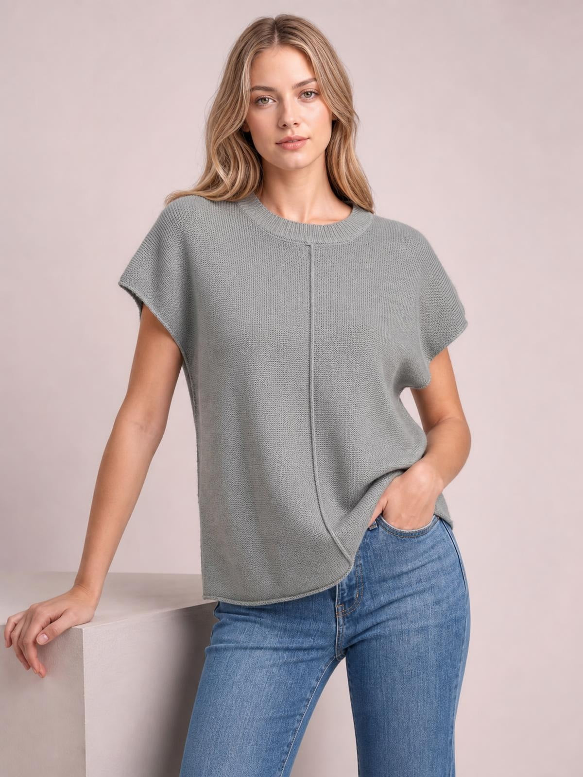 Model poses in a gray exposed-seam round neck short sleeve sweater with a relaxed, boxy silhouette, one hand in her jeans pocket.