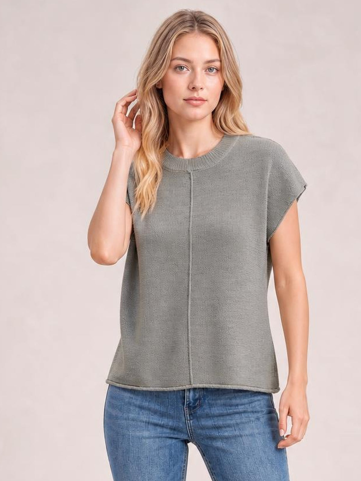 Model wearing a gray exposed seam round neck short sleeve sweater with a relaxed straight silhouette and ribbed neckline, posing with one hand in her hair.