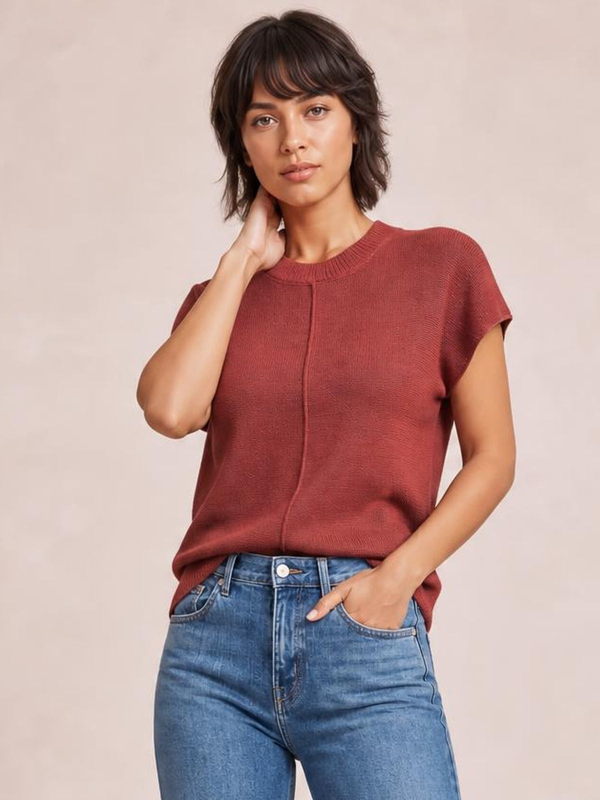 Model wears a brick-red exposed seam round neck short sleeve sweater with a relaxed straight silhouette, ribbed trim, and a hand resting at her neck.