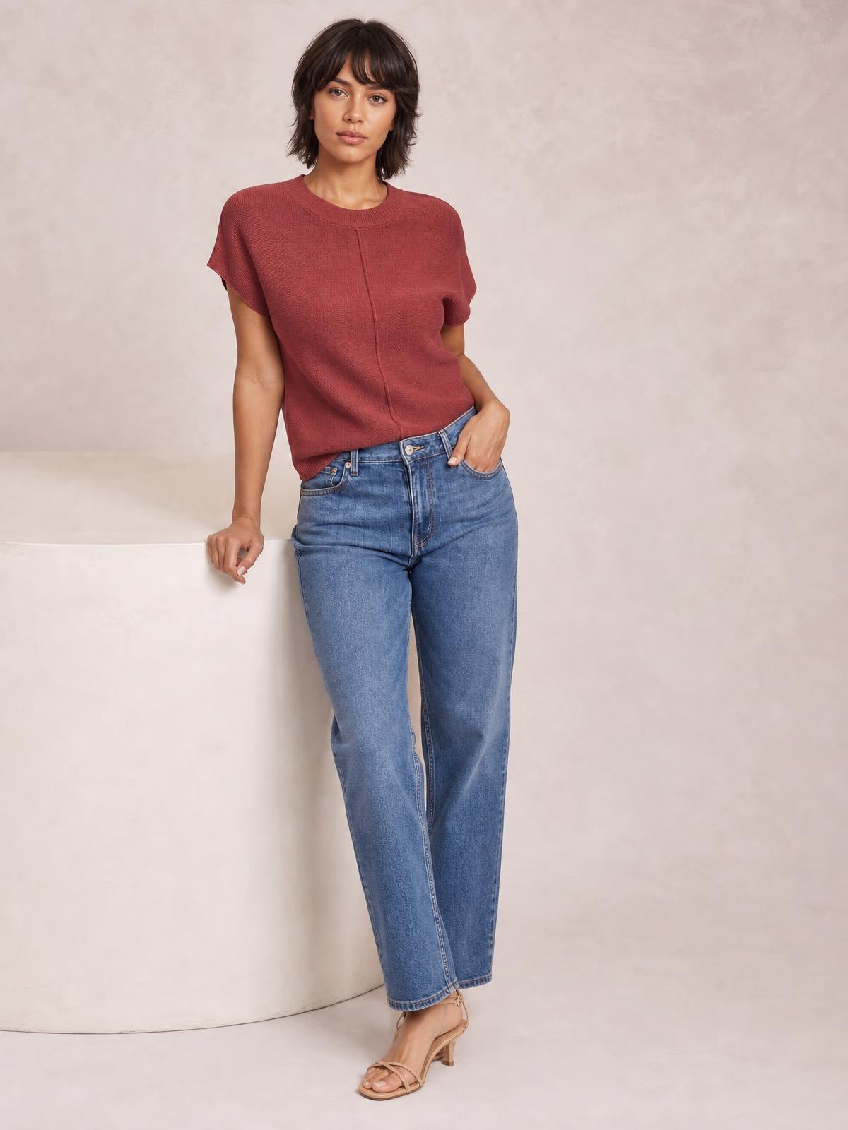 Model wearing a rust-red exposed seam sweater with a round neck, short sleeves, and relaxed silhouette, paired with straight-leg blue jeans.