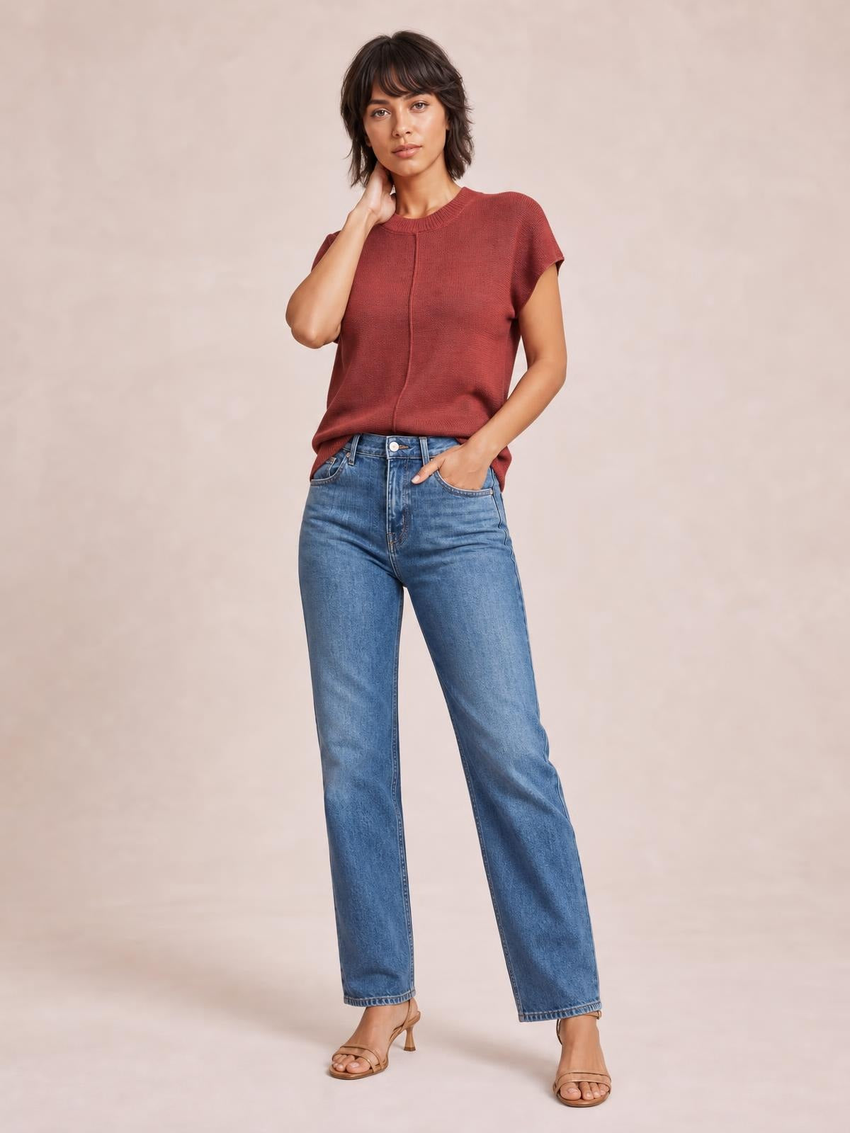 A model wears a rust-red round-neck short-sleeve sweater with an exposed center seam and relaxed silhouette, posing with one hand at her neck and one in her pocket.