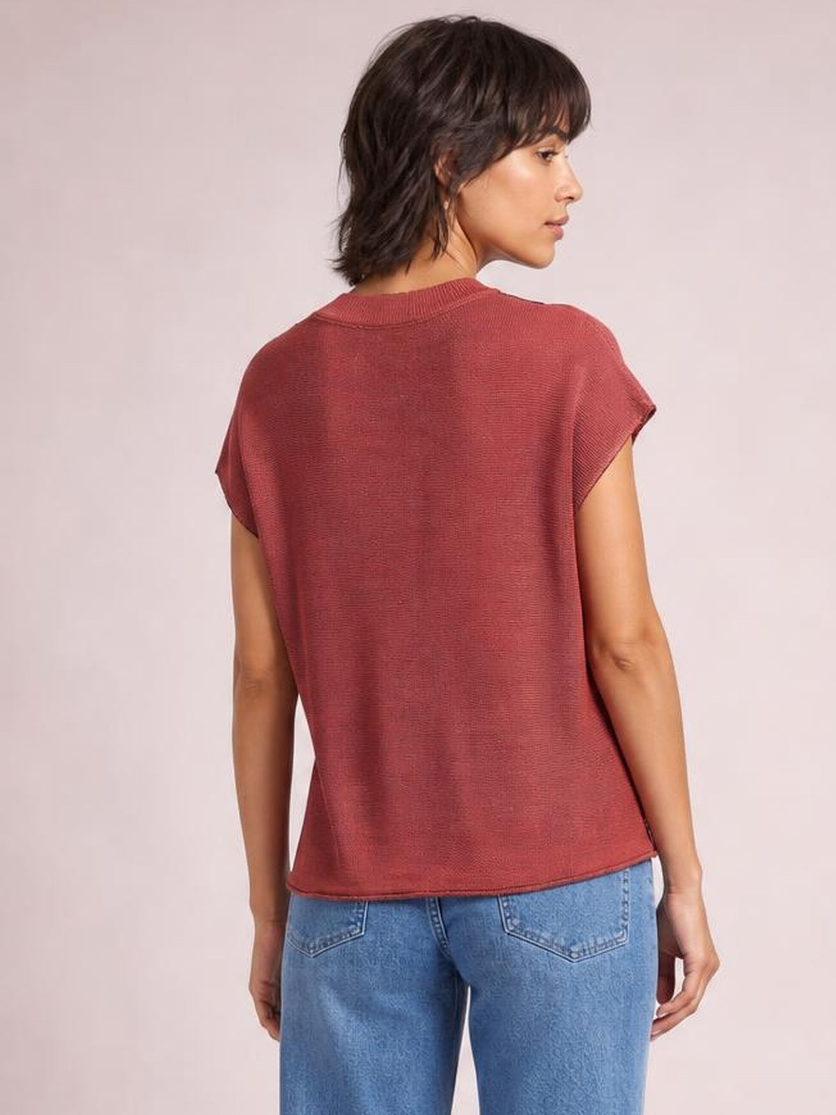 Model shown from the back in a rust-red round-neck short-sleeve sweater with a relaxed straight silhouette and visible exposed seam detailing, turning her head aside.