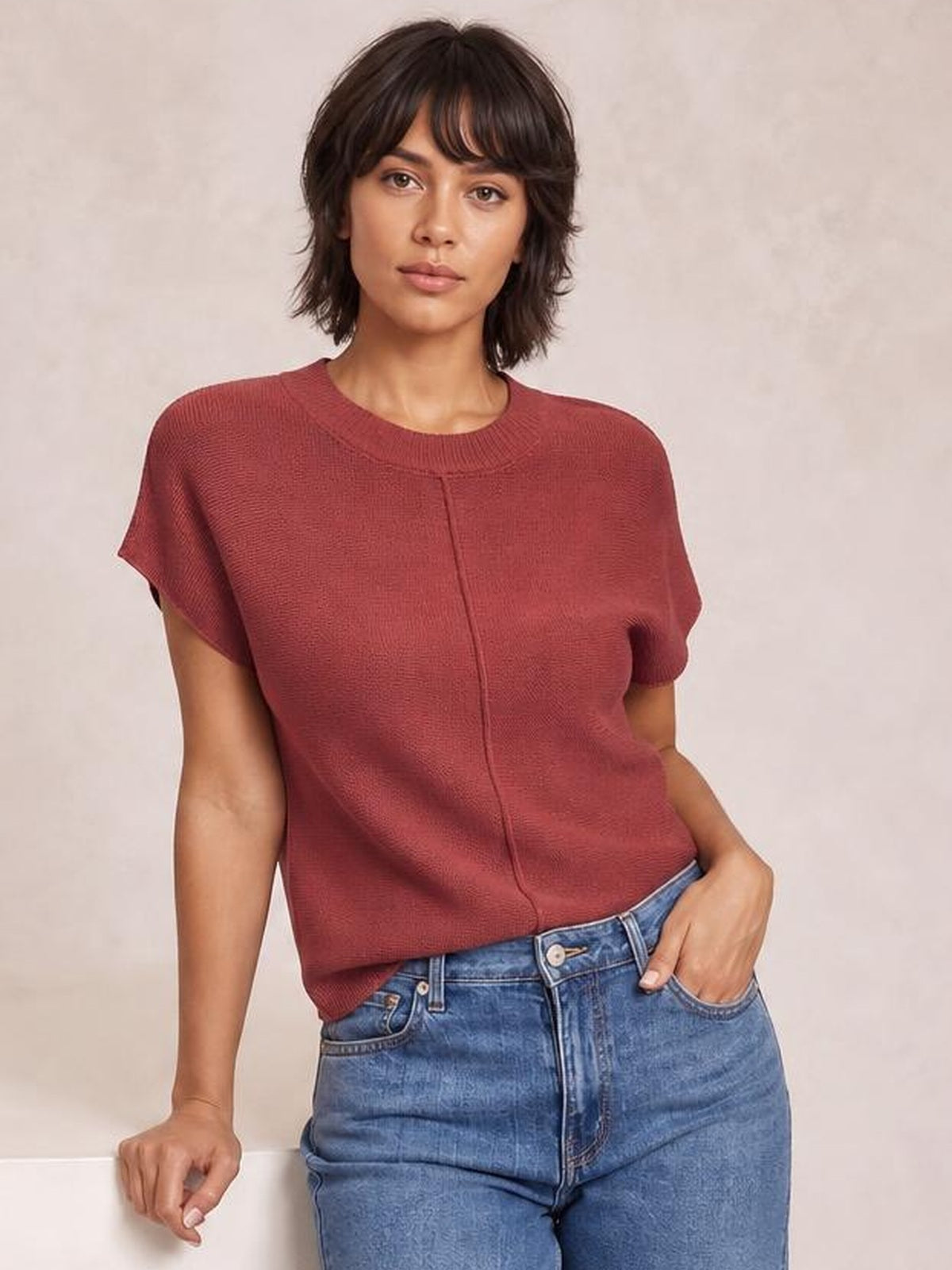 Model wearing a burgundy exposed-seam round-neck short-sleeve sweater with a relaxed silhouette, leaning casually with one hand in her jeans pocket.