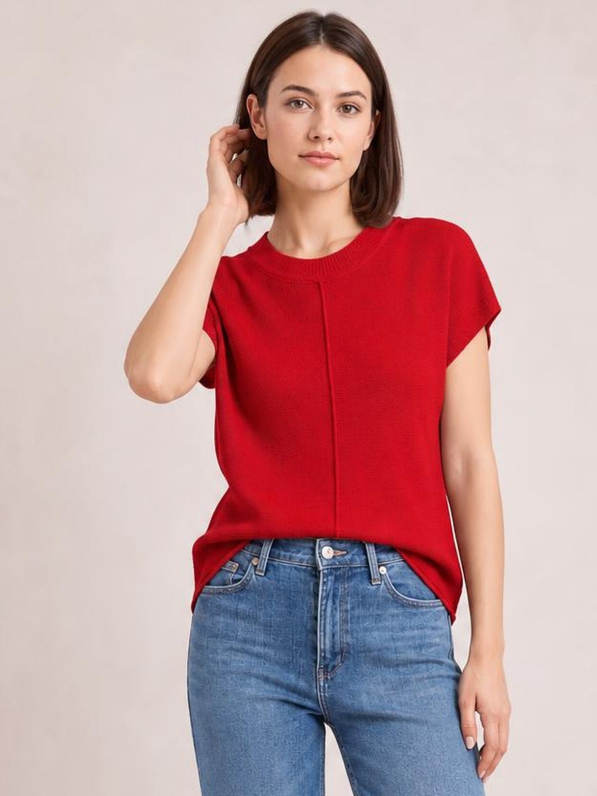 A model wears a red round-neck short-sleeve sweater with an exposed center seam and relaxed silhouette, posing with one hand touching her hair.