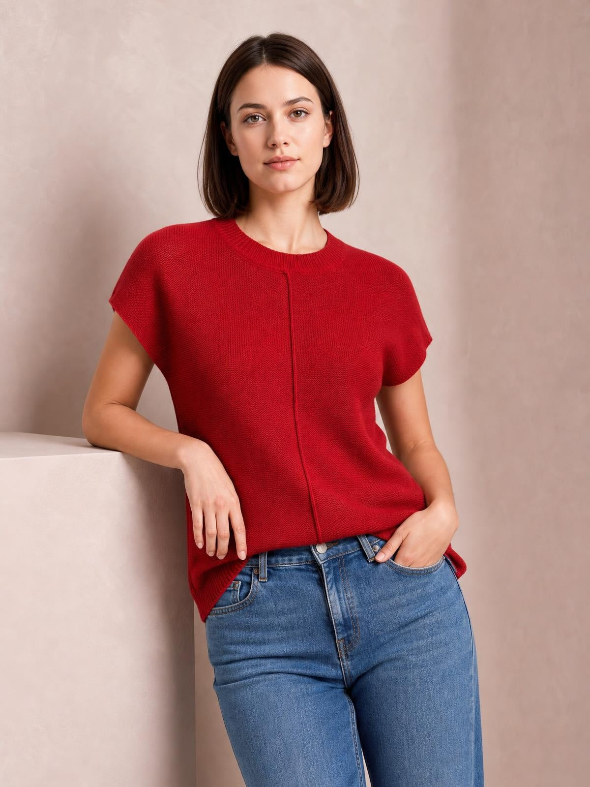 A model poses in a red exposed-seam sweater with a round neck, short sleeves, and a relaxed silhouette, paired with blue jeans.