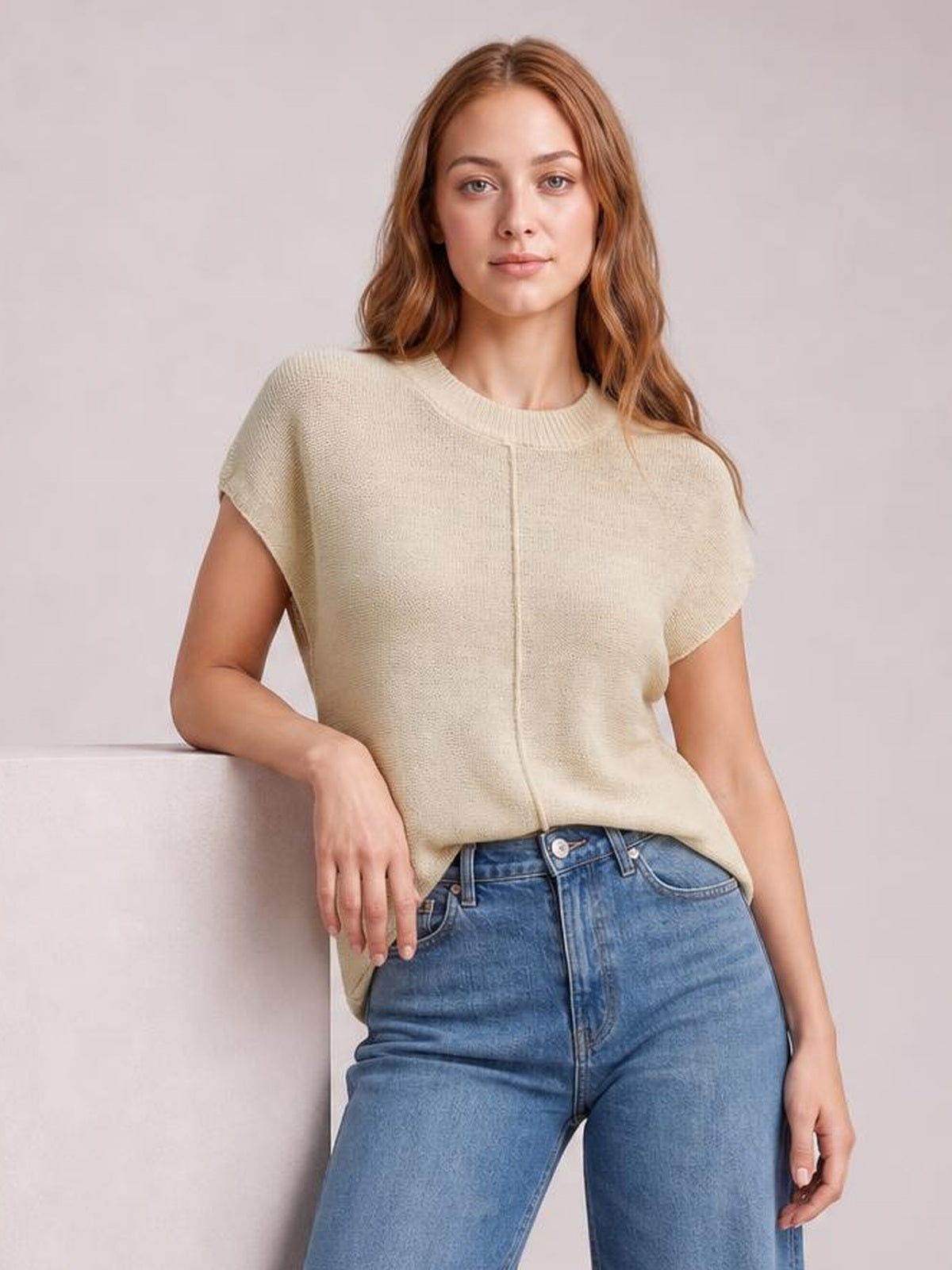 Model wears a cream exposed-seam round-neck short-sleeve sweater with a relaxed silhouette and subtle knit texture, posed with one hand resting on a ledge.