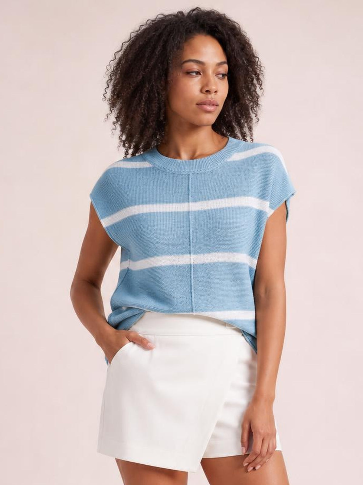 A model wears a light blue striped knit top with a round neck, exposed center seam, short sleeves, and a relaxed silhouette, standing with one hand in her shorts pocket.