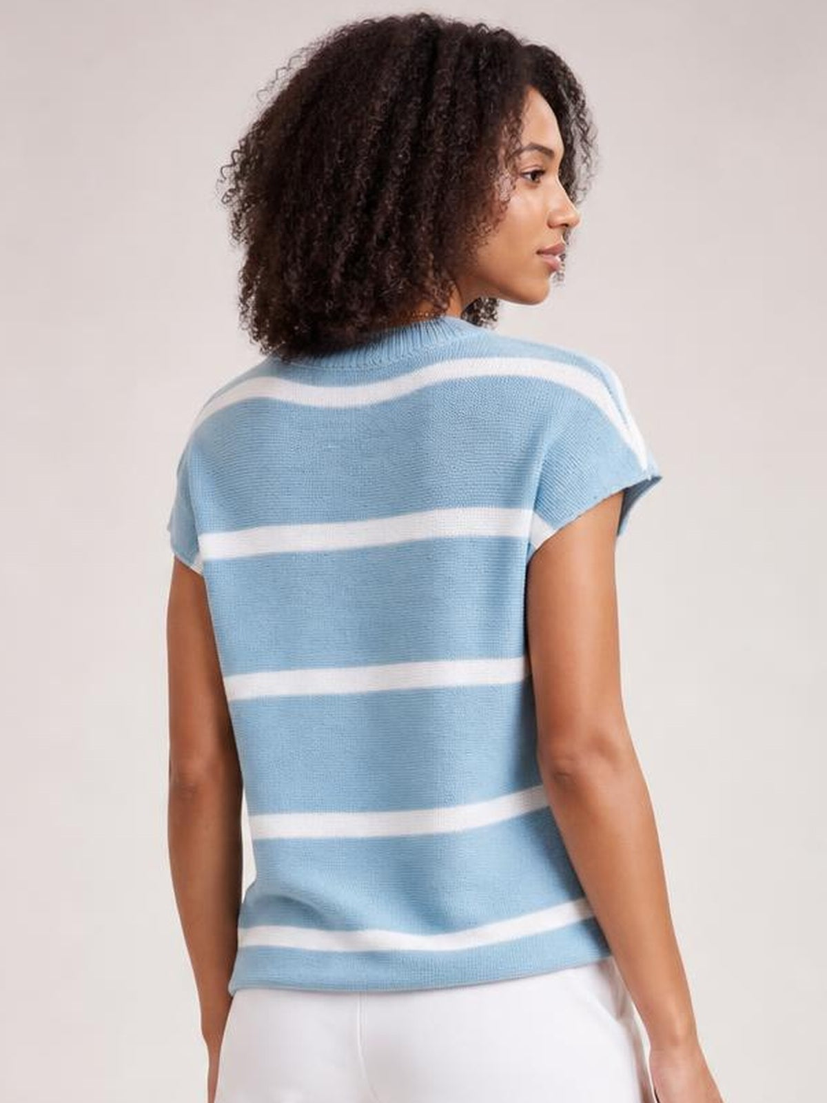 A model turned away wears a light blue striped knit top with white horizontal bands, round neck, short sleeves, and a relaxed silhouette.