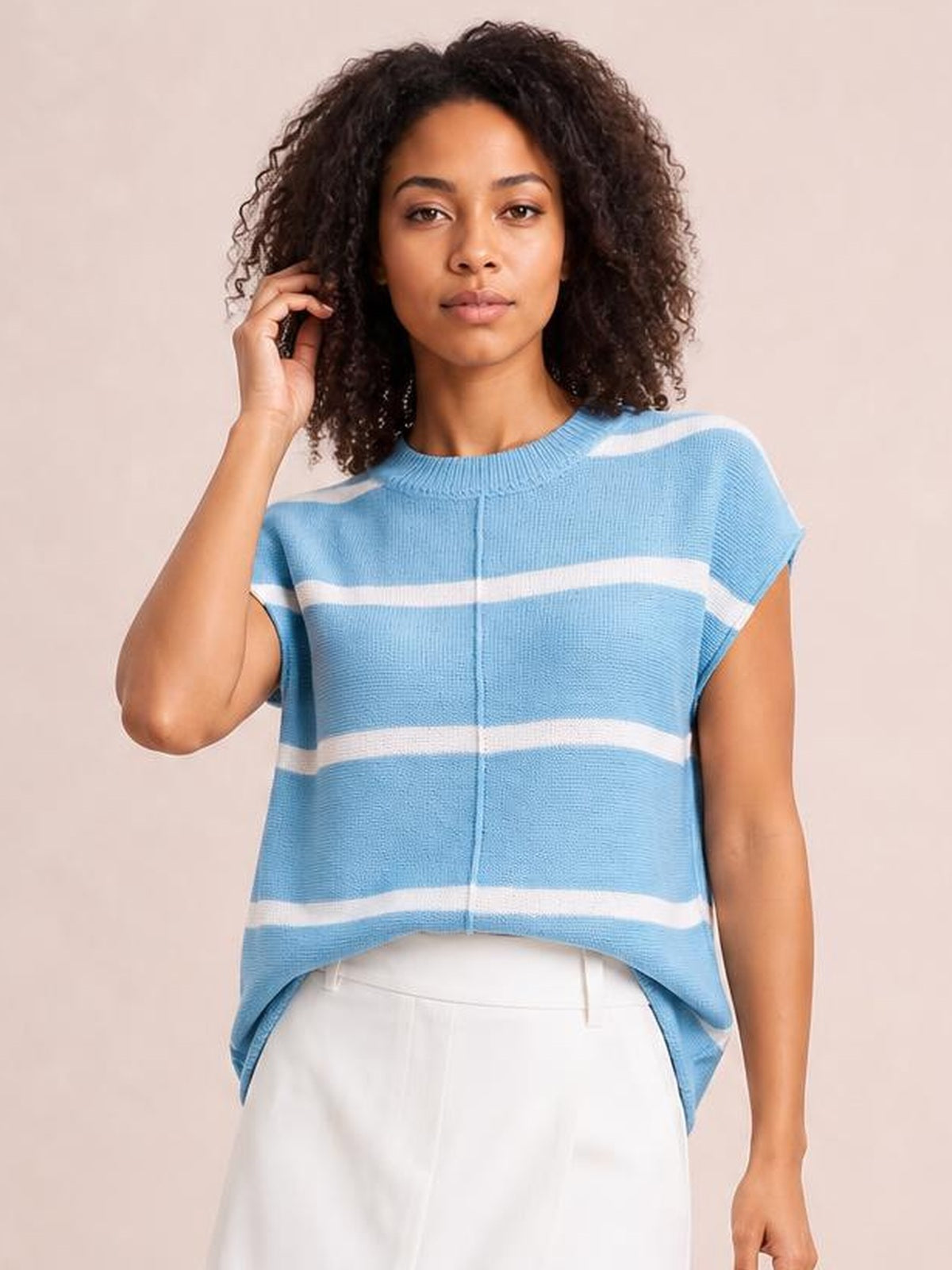 A model touches her hair while wearing a light blue and white striped knit top with a round neck, exposed center seam, short sleeves, and a relaxed curved hem.