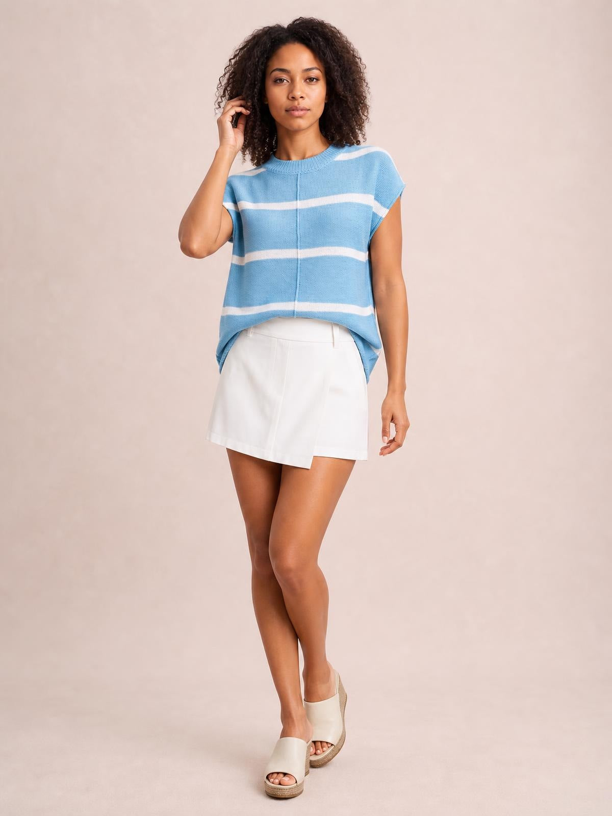 Model poses in a blue and white striped round neck knit top with exposed center seam, relaxed boxy silhouette, short sleeves, and curved hem.