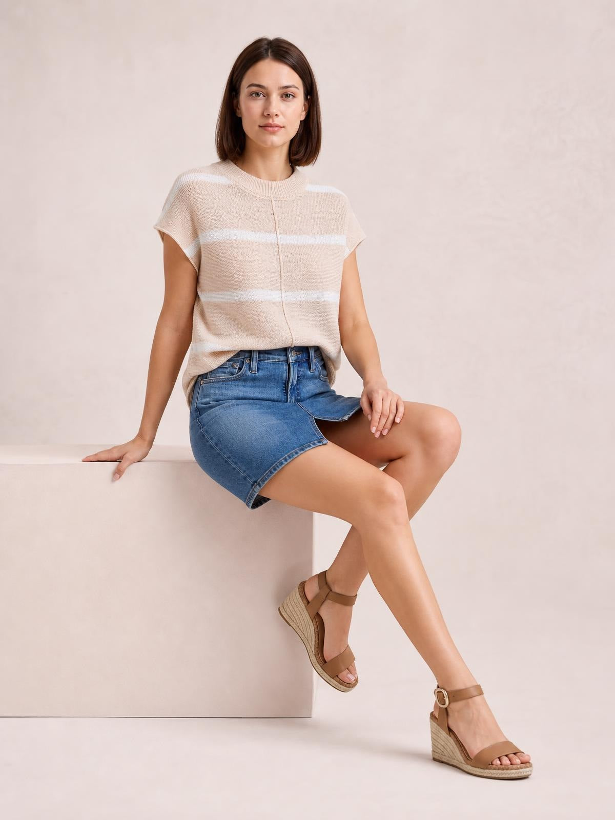 Model seated in a beige knit top with white horizontal stripes, a round neck, exposed center seam, short sleeves, and a relaxed silhouette.