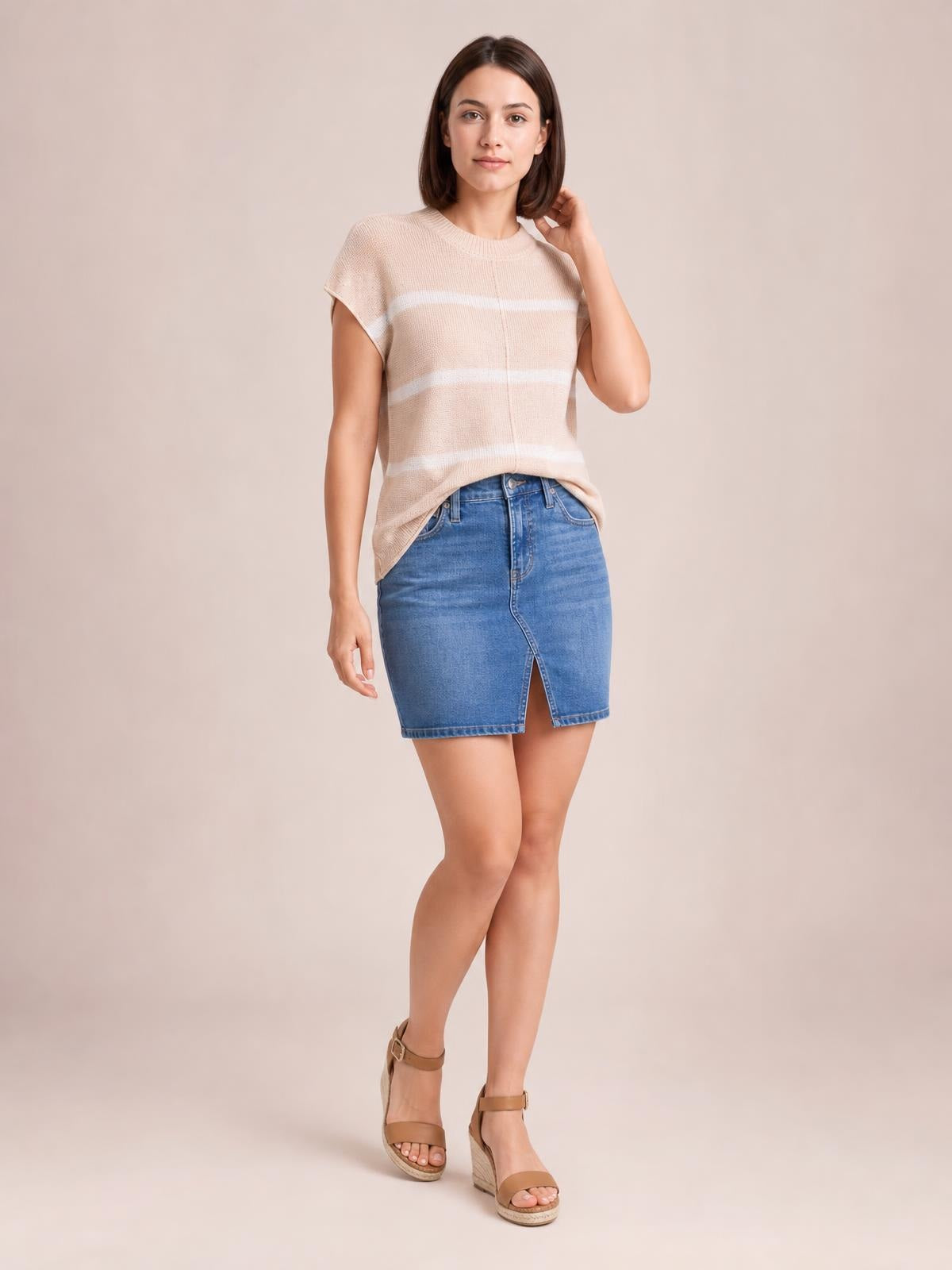 A model wears a beige striped round-neck knit top with exposed center seam and short sleeves, posing with one hand in her hair and crossed legs.