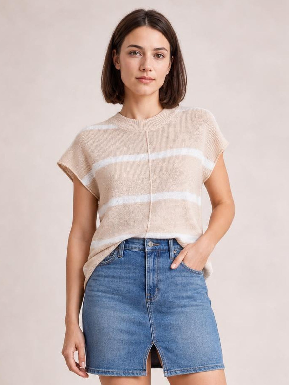A model wears a beige and white striped round-neck knit top with exposed center seam, short sleeves, and a relaxed silhouette, posing with one hand in her pocket.