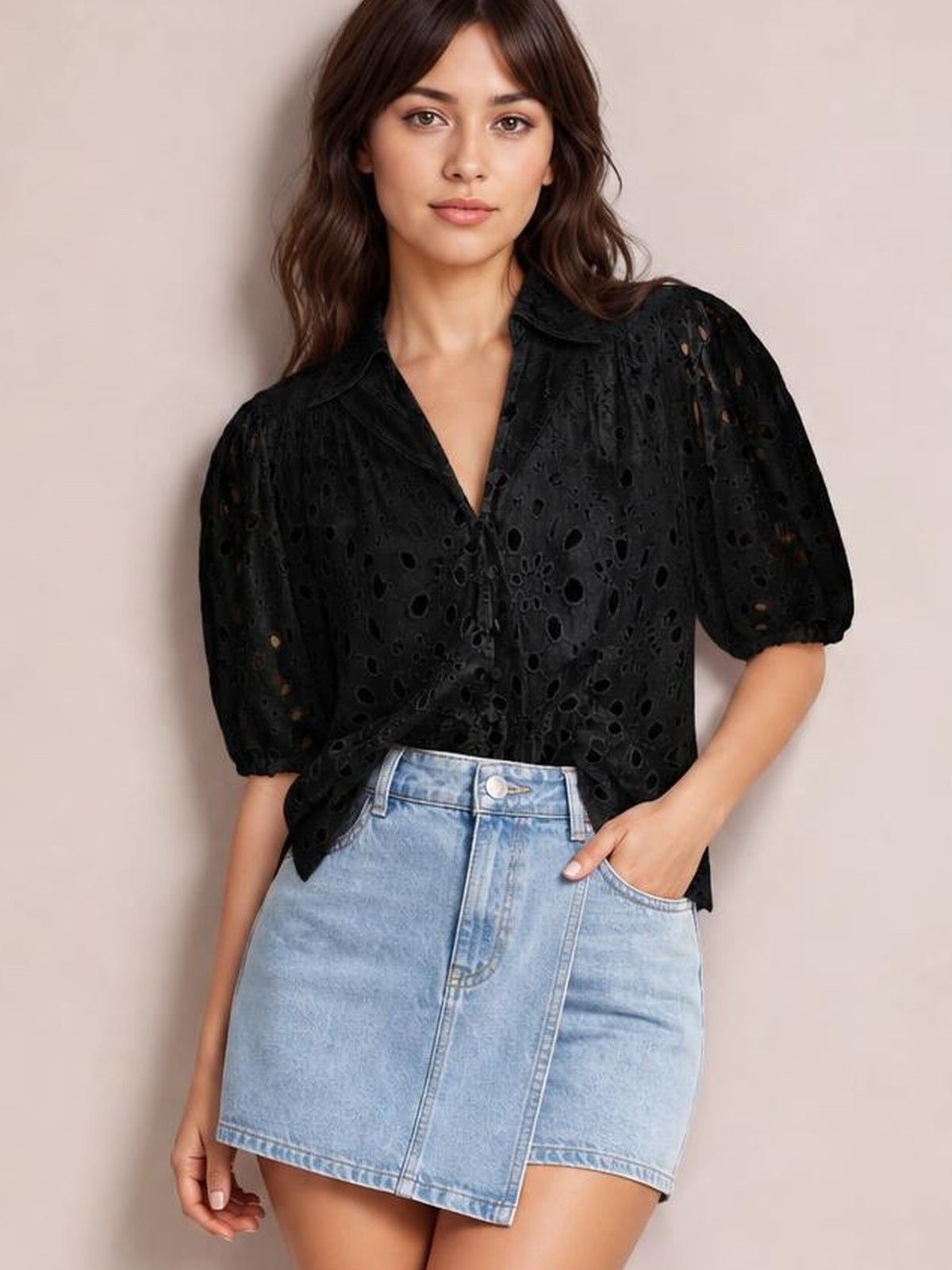 Model wears a black eyelet button-up blouse with a collared V-neck, puffed half sleeves, and a relaxed silhouette tucked into a denim skirt.