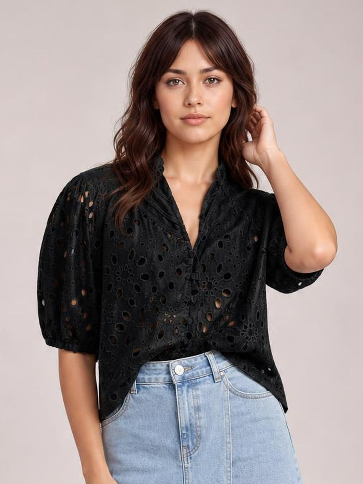 Model wearing a black eyelet button-up blouse with half sleeves, an airy relaxed silhouette, and an open V-neckline, posing with one hand in her hair.