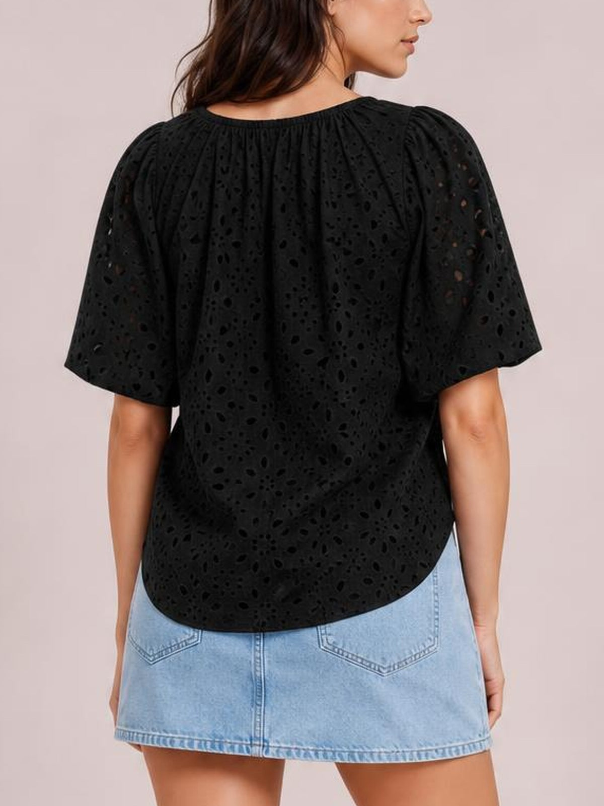 A model wears a black eyelet blouse with gathered neckline, loose silhouette, and half sleeves, shown from the back with a curved hem over a denim skirt.