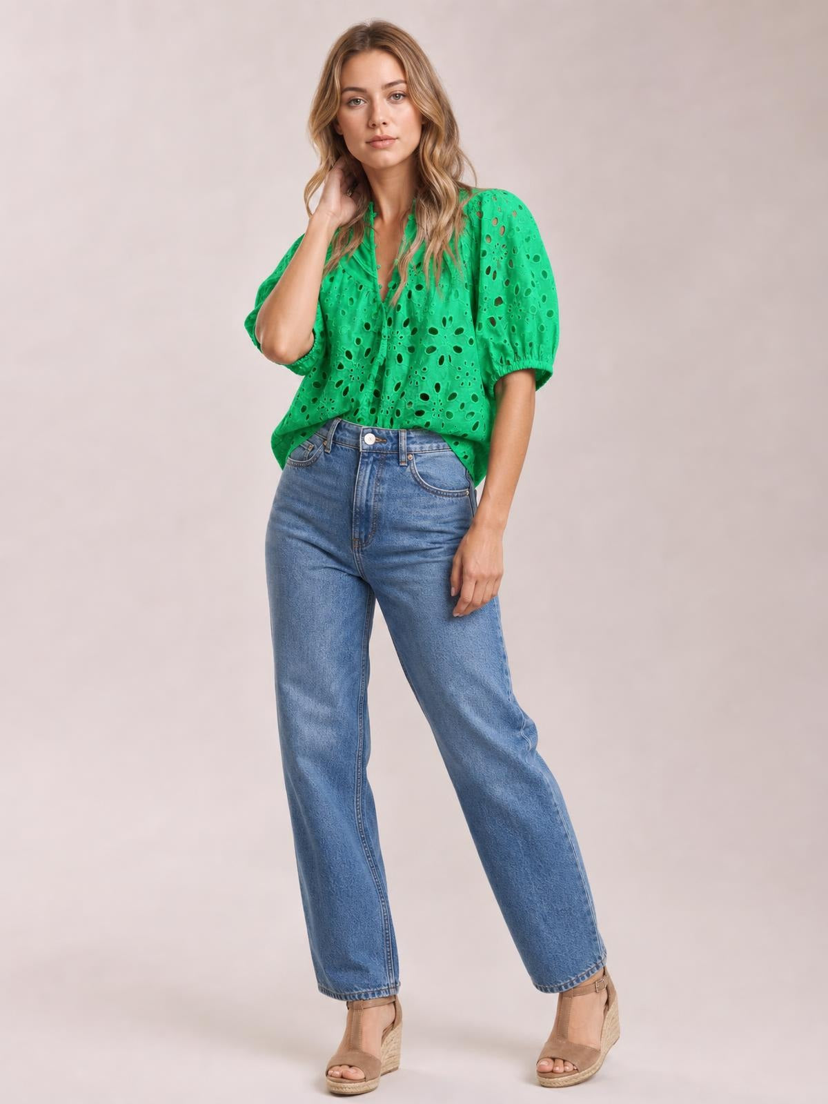 A model wears a green eyelet button-up blouse with puffed half sleeves, a relaxed silhouette, and a gathered neckline, posing with one hand touching her hair.