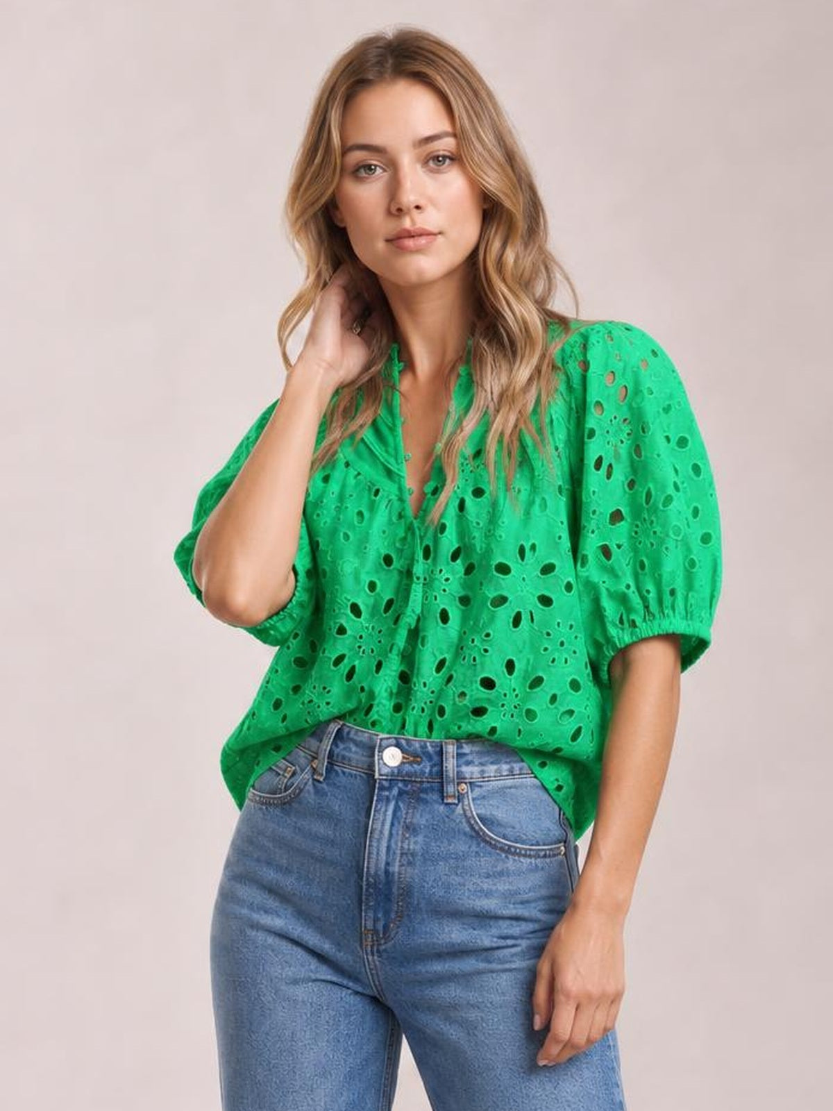 A model poses with one hand in her hair wearing a bright green eyelet button-up blouse with half puff sleeves, visible buttons, and a relaxed silhouette.