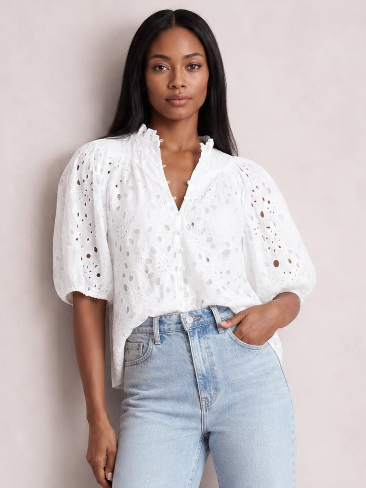 A model wears a white eyelet button-up blouse with a ruffled neckline, puffed half sleeves, and a relaxed silhouette, posing with one hand in her jeans pocket.