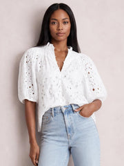 A model wears a white eyelet button-up blouse with a ruffled neckline, puffed half sleeves, and a relaxed silhouette, posing with one hand in her jeans pocket.