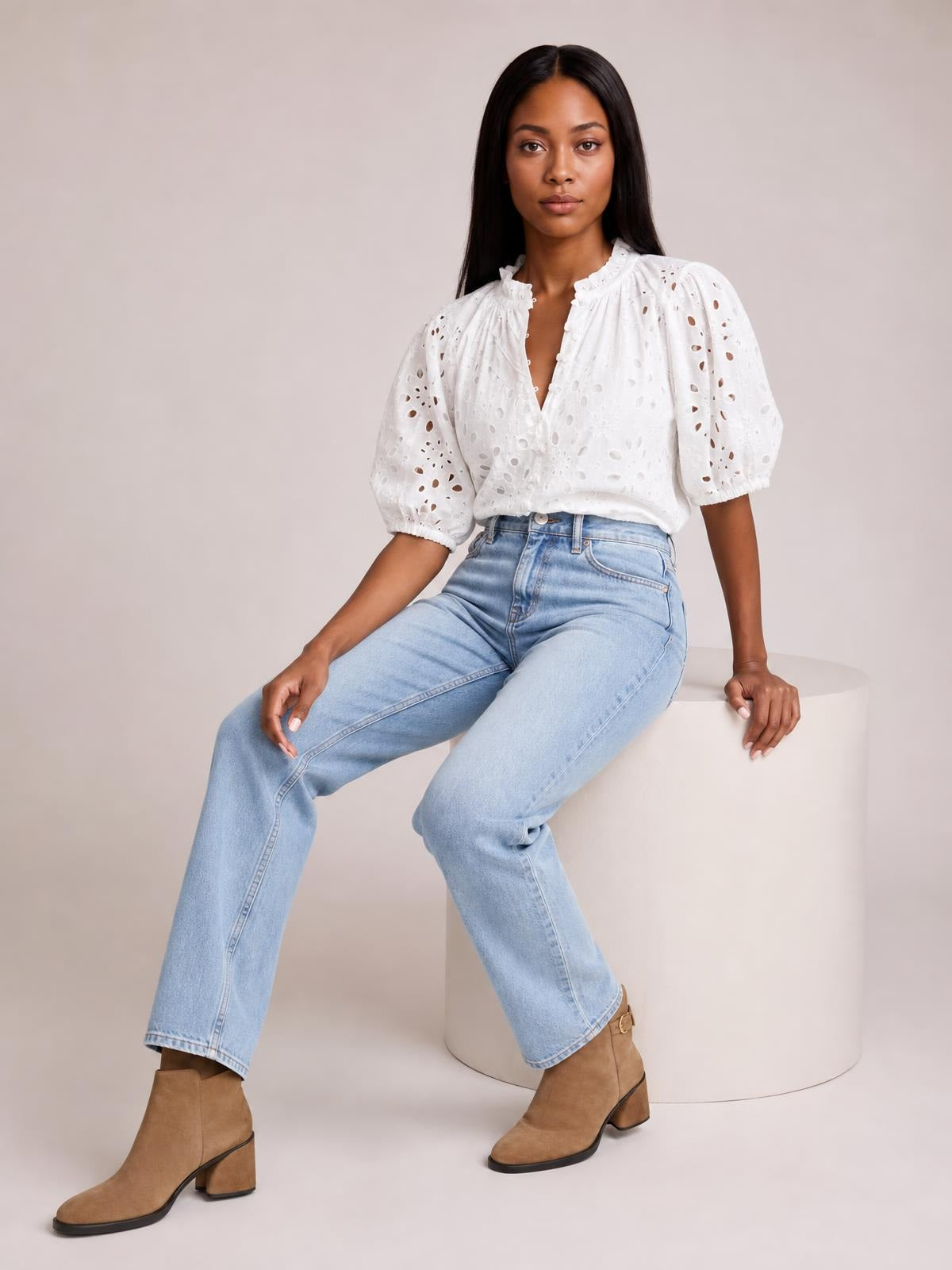 A model sits wearing a white eyelet button-up blouse with a gathered neckline, half sleeves, elastic cuffs, and a relaxed silhouette tucked into light-wash jeans.