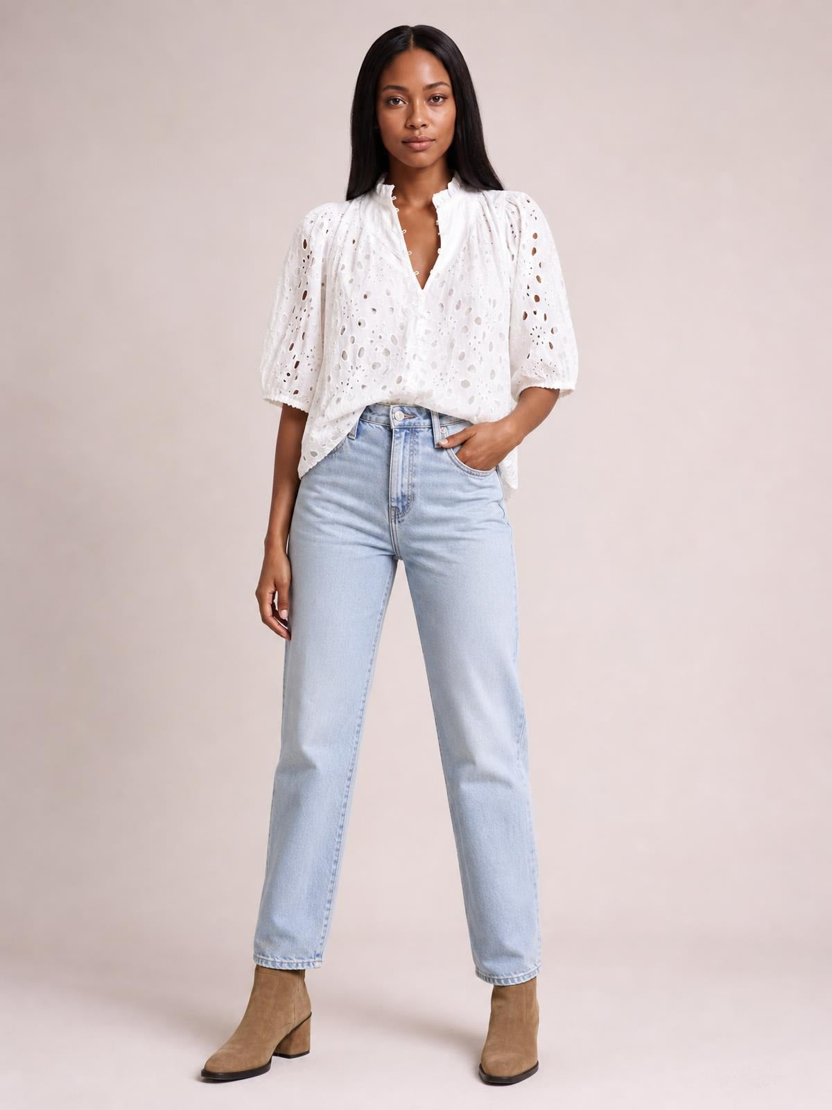 Model poses in a white eyelet button-up blouse with a relaxed silhouette, split neckline, and half sleeves, paired with light-wash jeans.