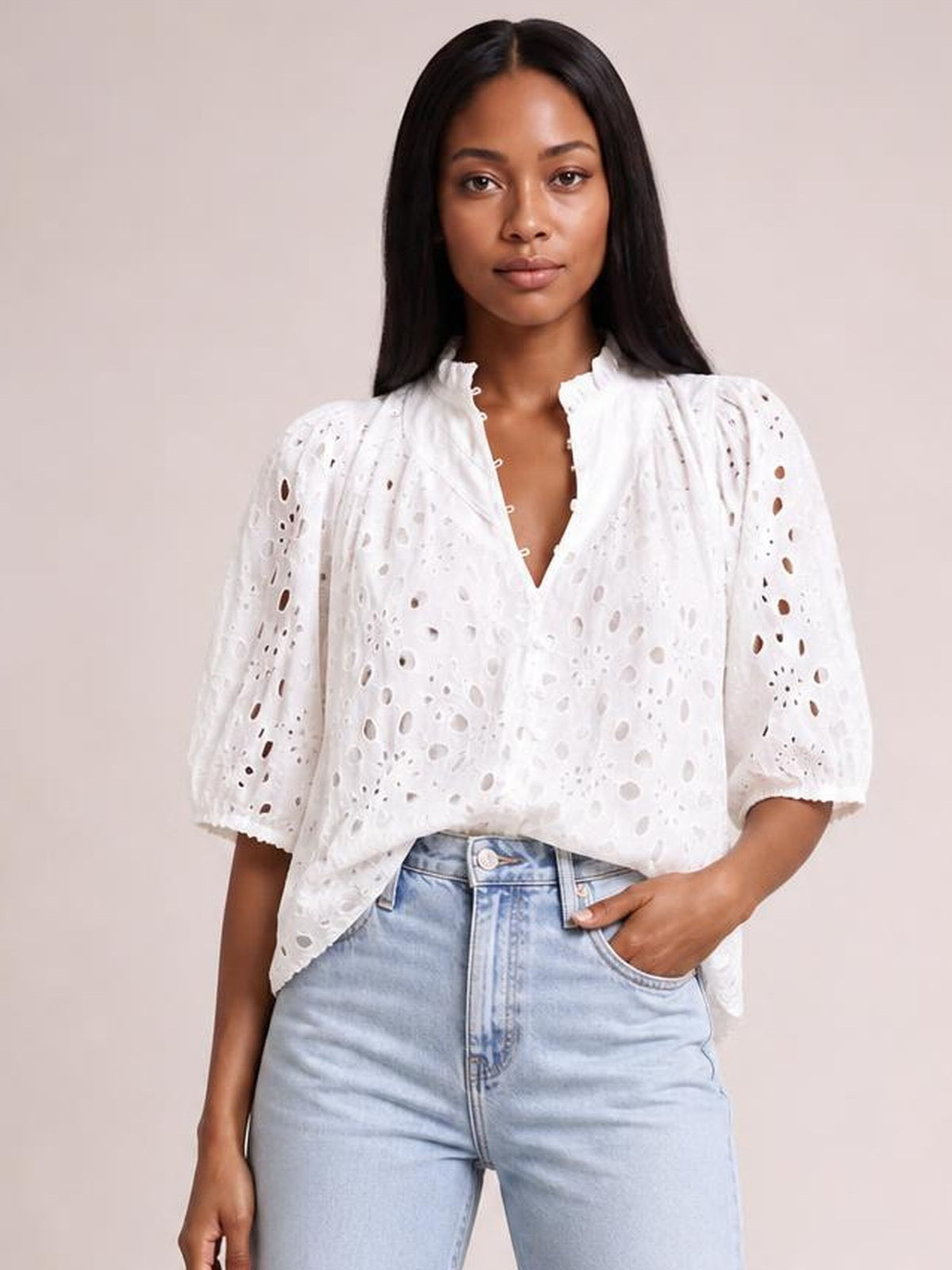 Model poses with one hand in her pocket, wearing a white eyelet blouse with a button-up neckline, ruffled collar, puffed half sleeves, and relaxed silhouette.