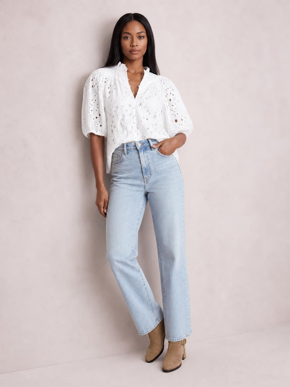A model stands with one hand in her pocket wearing a white opaque eyelet button-up blouse with half sleeves, a relaxed silhouette, and gathered shoulders.