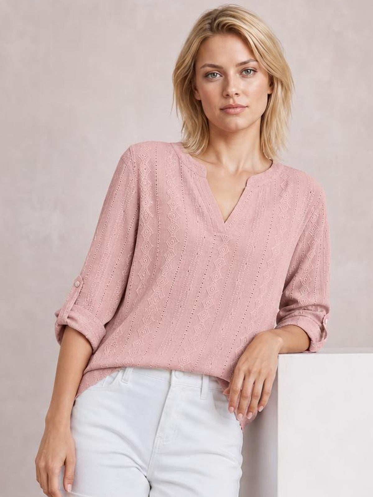 Model wears a dusty pink eyelet top with a notched neckline, relaxed silhouette, and three-quarter roll-tab sleeves while leaning casually.