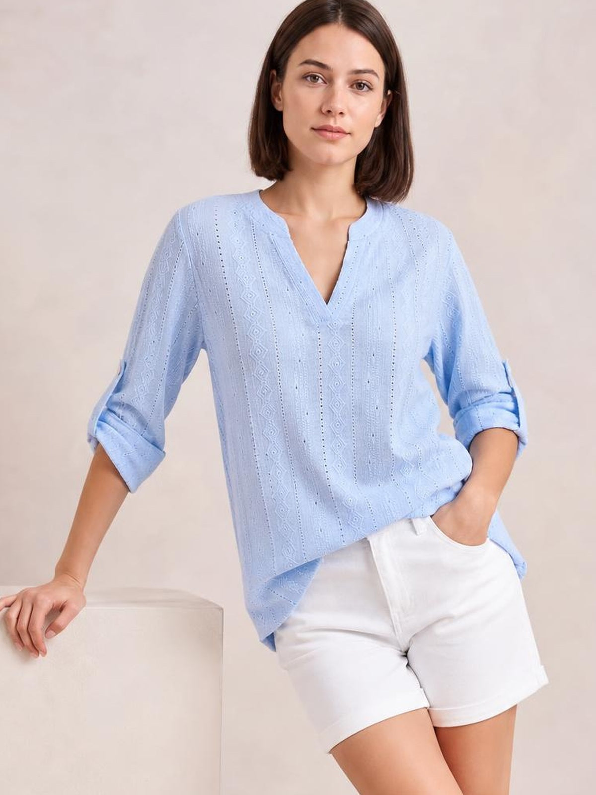 A model poses in a light blue eyelet top with a notched neckline, relaxed silhouette, and rolled three-quarter sleeves secured with tabs.