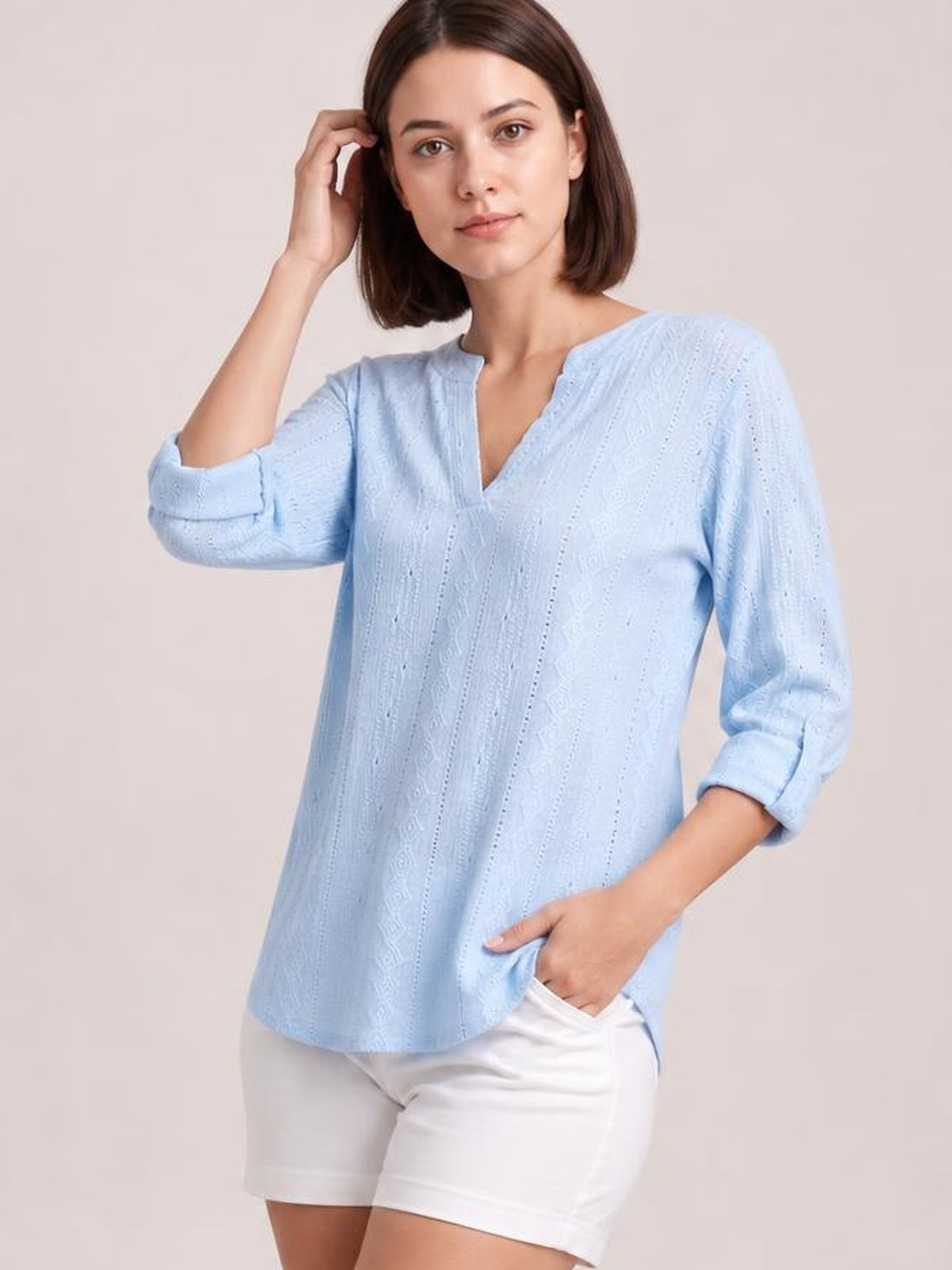 Model wearing a light blue eyelet top with a notched neckline, relaxed silhouette, and three-quarter roll-tab sleeves, posing with one hand in her shorts pocket.