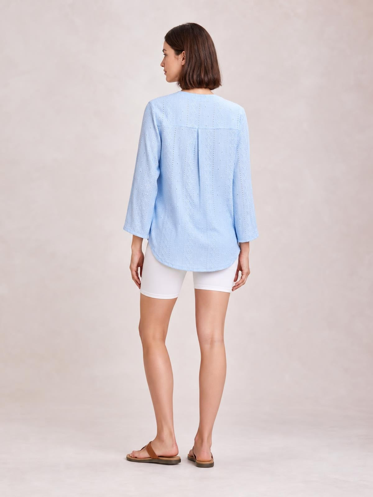 Model shown from behind wearing a light blue eyelet top with three-quarter sleeves, a relaxed silhouette, back yoke, center pleat, and curved hem.