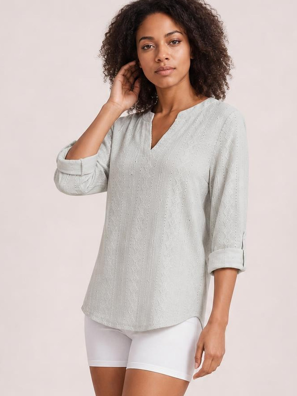 Model wearing a light gray eyelet top with a notched neckline, relaxed silhouette, and three-quarter roll-tab sleeves, posing with one hand near her hair.