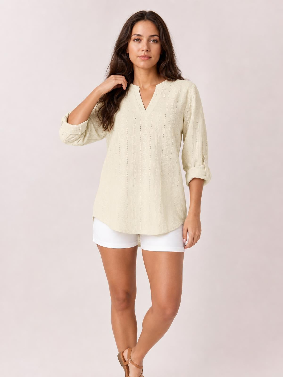 Model wears a cream eyelet top with a notched neckline, relaxed straight silhouette, and long roll-tab sleeves, posing with one hand in her hair.