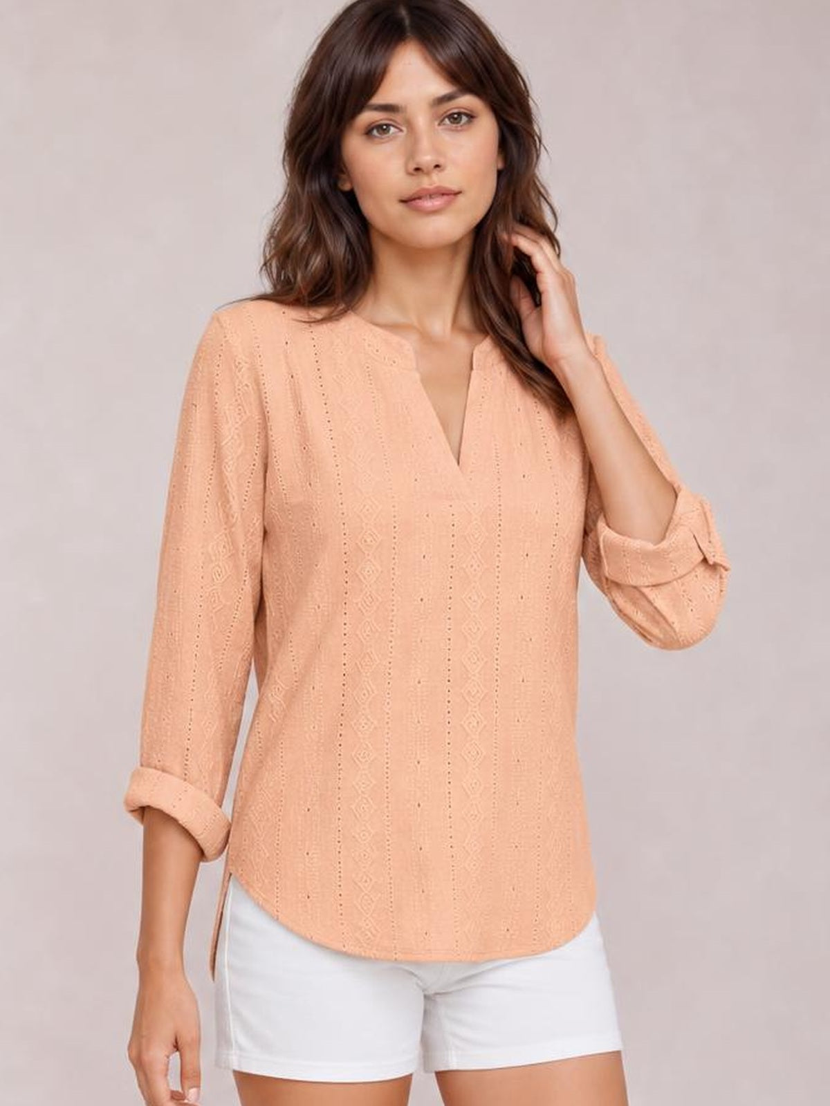 A model wears a peach eyelet top with a notched V-neckline, relaxed straight silhouette, and rolled long sleeves secured with tabs, posing with one hand in her hair.