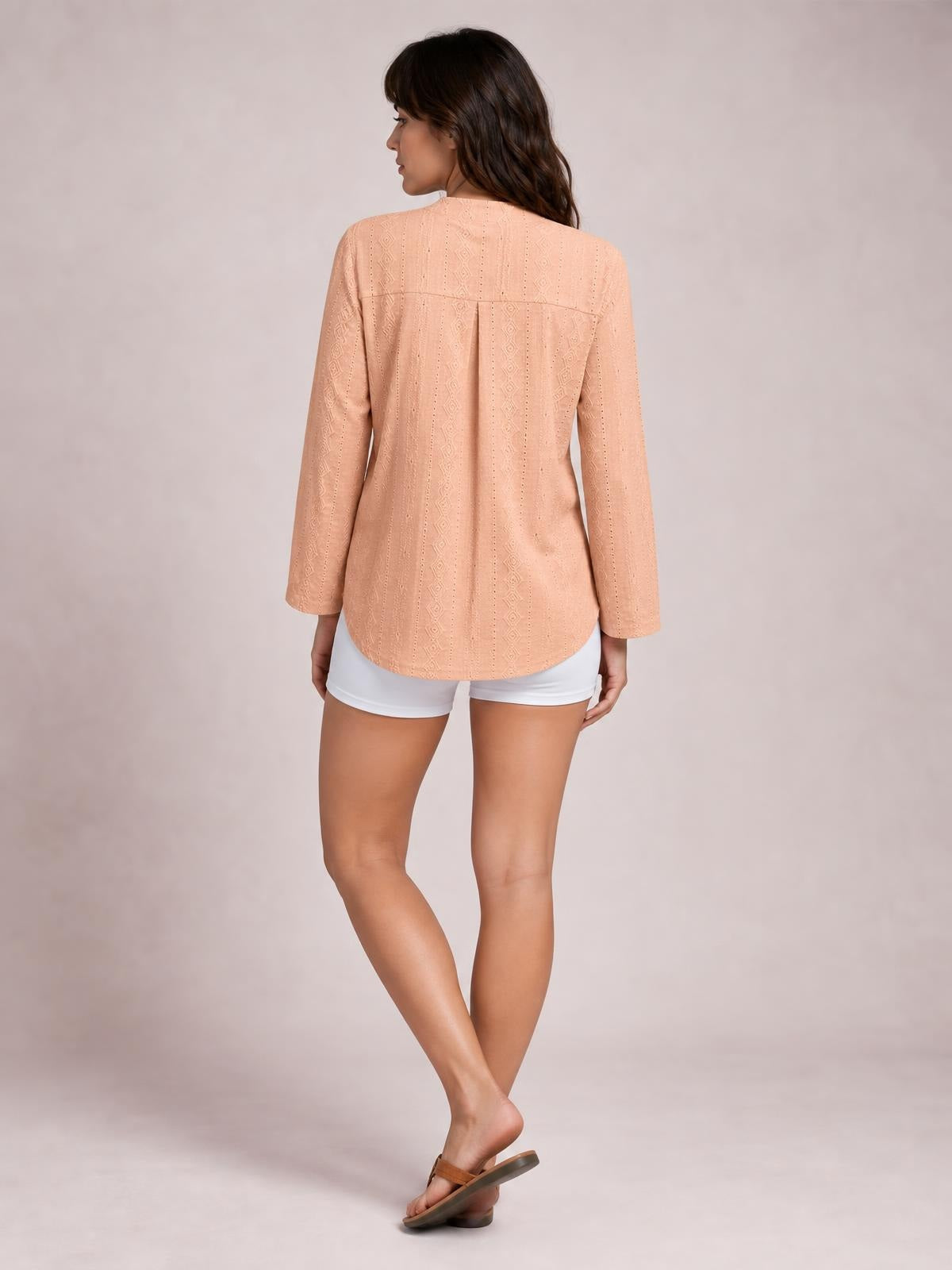A model stands with crossed legs, wearing a peach eyelet top with long sleeves, a relaxed silhouette, back yoke, center pleat, and rounded hem.