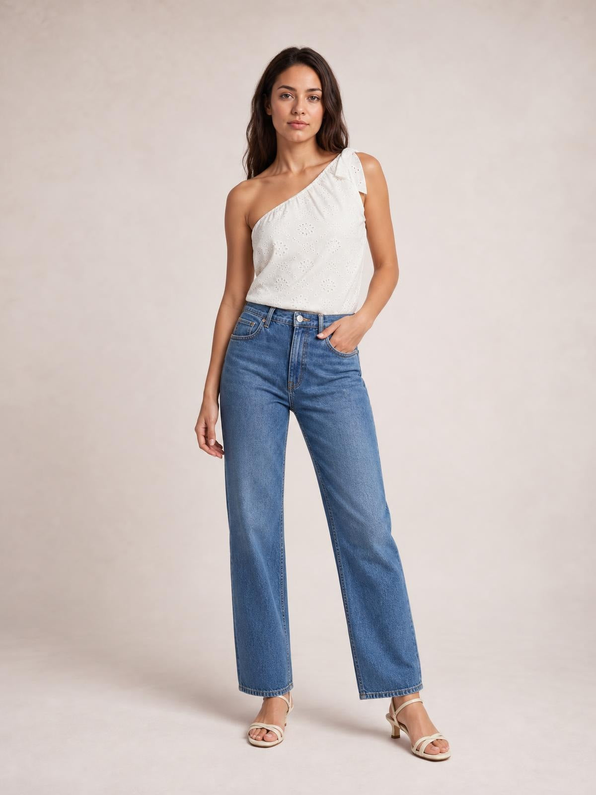 A model wears a white opaque eyelet one-shoulder tank with a tied shoulder detail and relaxed silhouette, standing with one hand in her jeans pocket.
