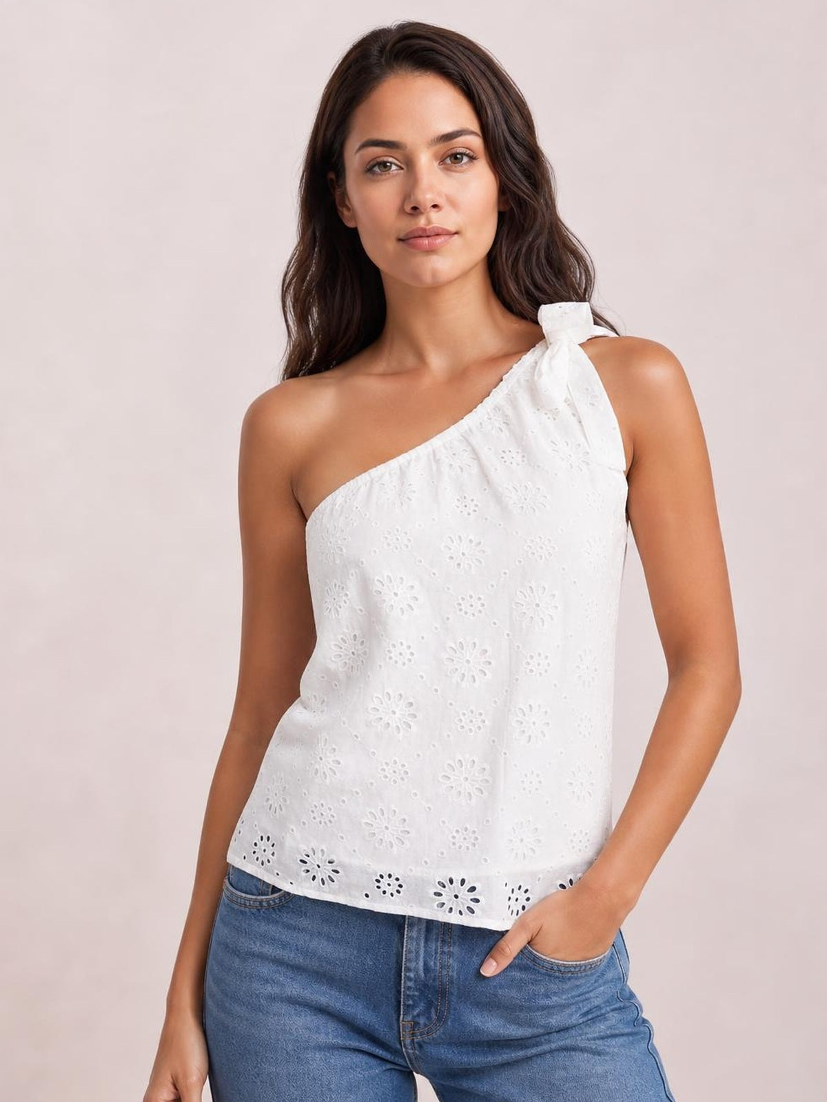 Model poses with one hand in a pocket, wearing a white eyelet one-shoulder tank with a tied shoulder detail and relaxed straight silhouette.