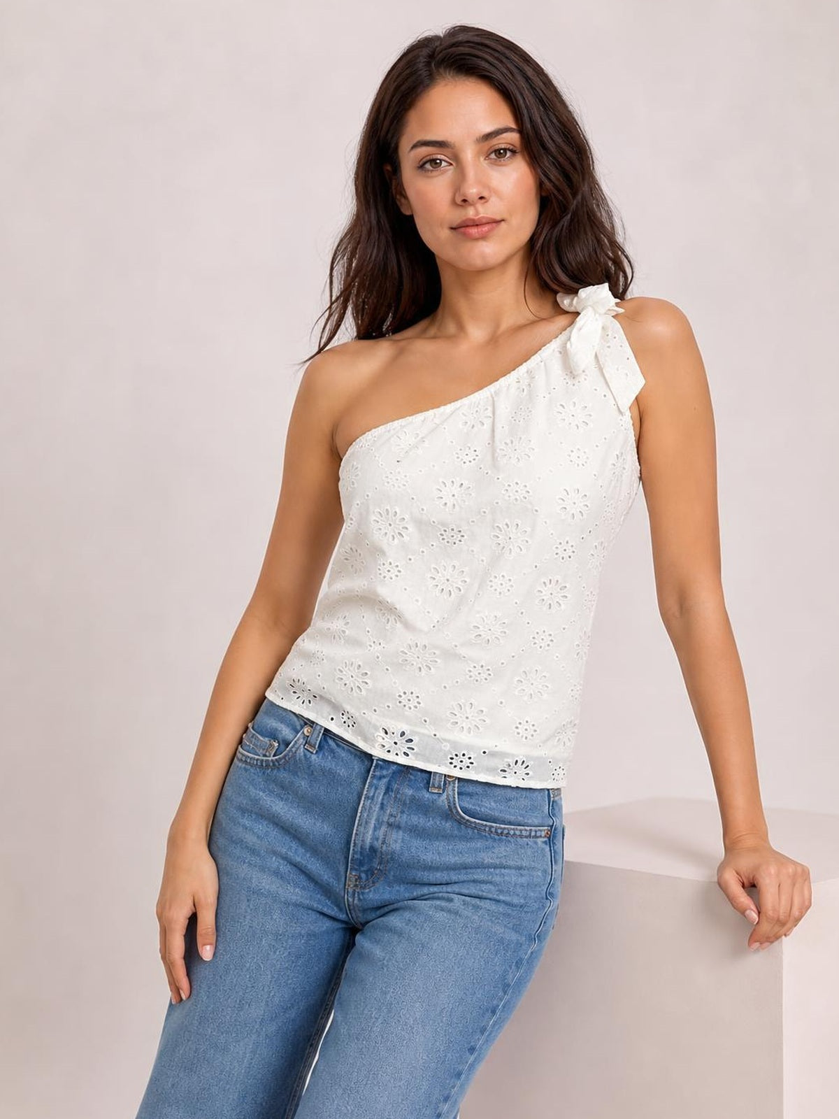 Model leans against a ledge wearing a white eyelet one-shoulder tank with a delicate shoulder tie and relaxed straight silhouette.