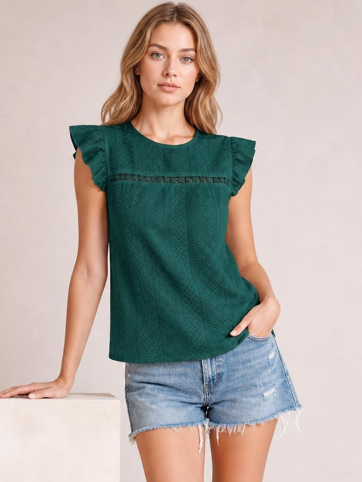 Model wearing a dark teal eyelet round neck cap sleeve top with cutout detailing and a relaxed straight silhouette, posing with one hand in her shorts pocket.