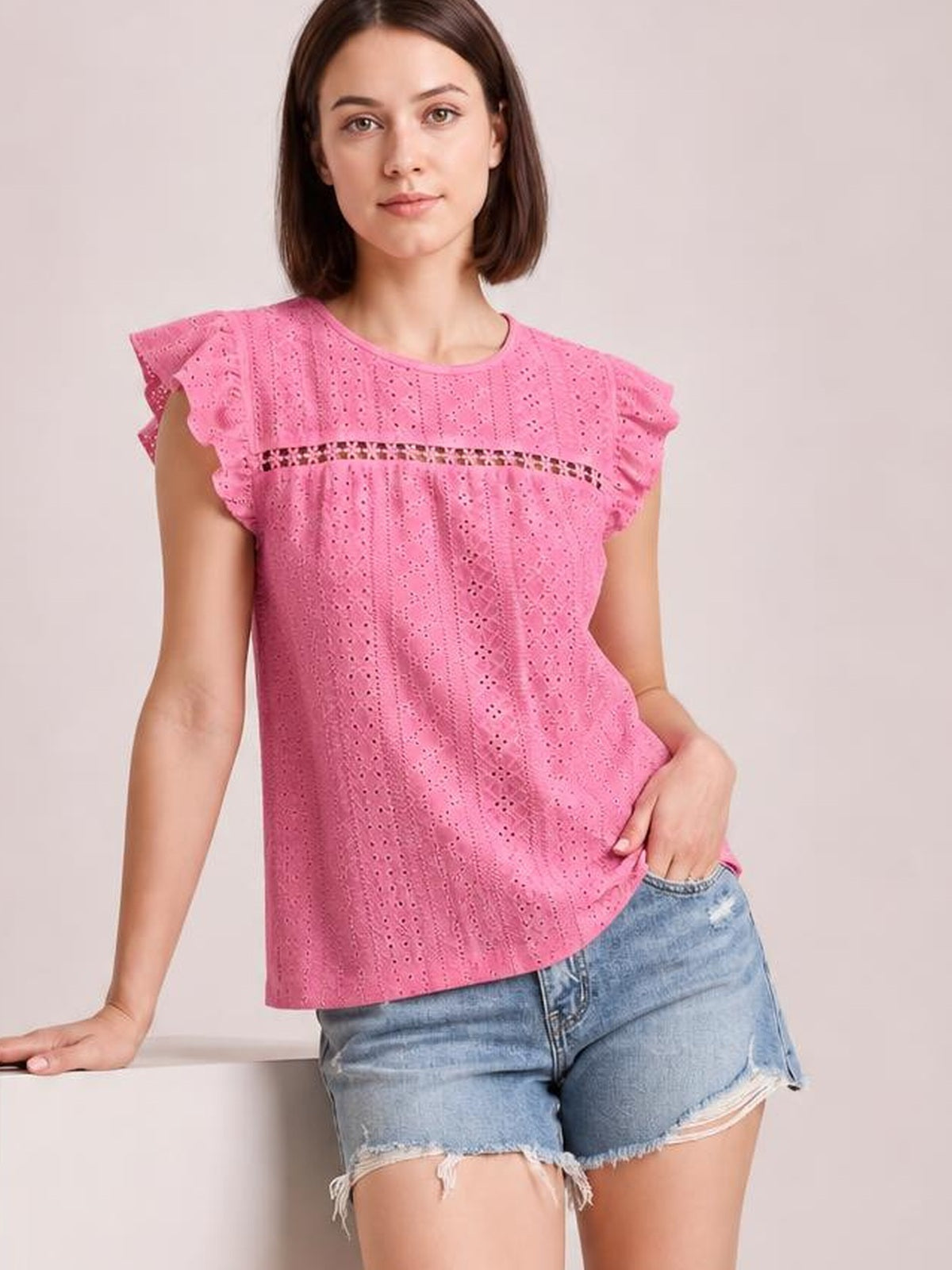 Model wearing a pink eyelet top with a round neck, ruffled cap sleeves, horizontal cutout panel, and relaxed straight silhouette, posing with one hand in her shorts pocket.
