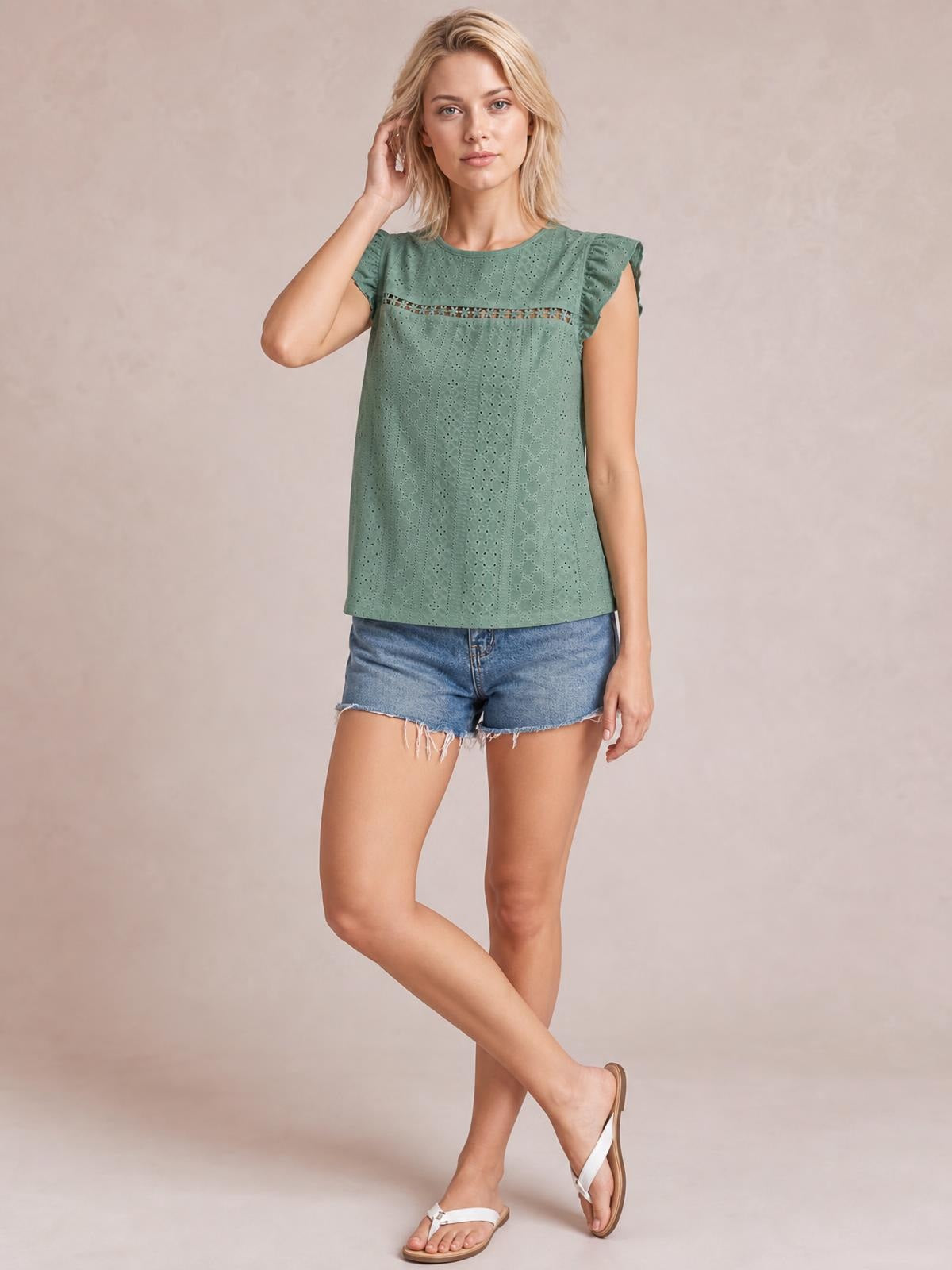 Model wears a sage green eyelet round-neck cap-sleeve top with decorative cutouts and a relaxed straight silhouette, posing with one hand in her hair.